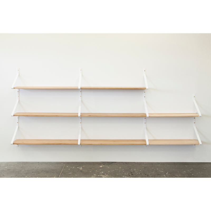 The Ribbon Shelf by One For Hundred is an elegant wall-mounted unit featuring four solid wood shelves, supported by white powder-coated steel brackets. Set against a plain white wall with a dark speckled floor, the evenly spaced shelves offer a sleek and graceful appearance.