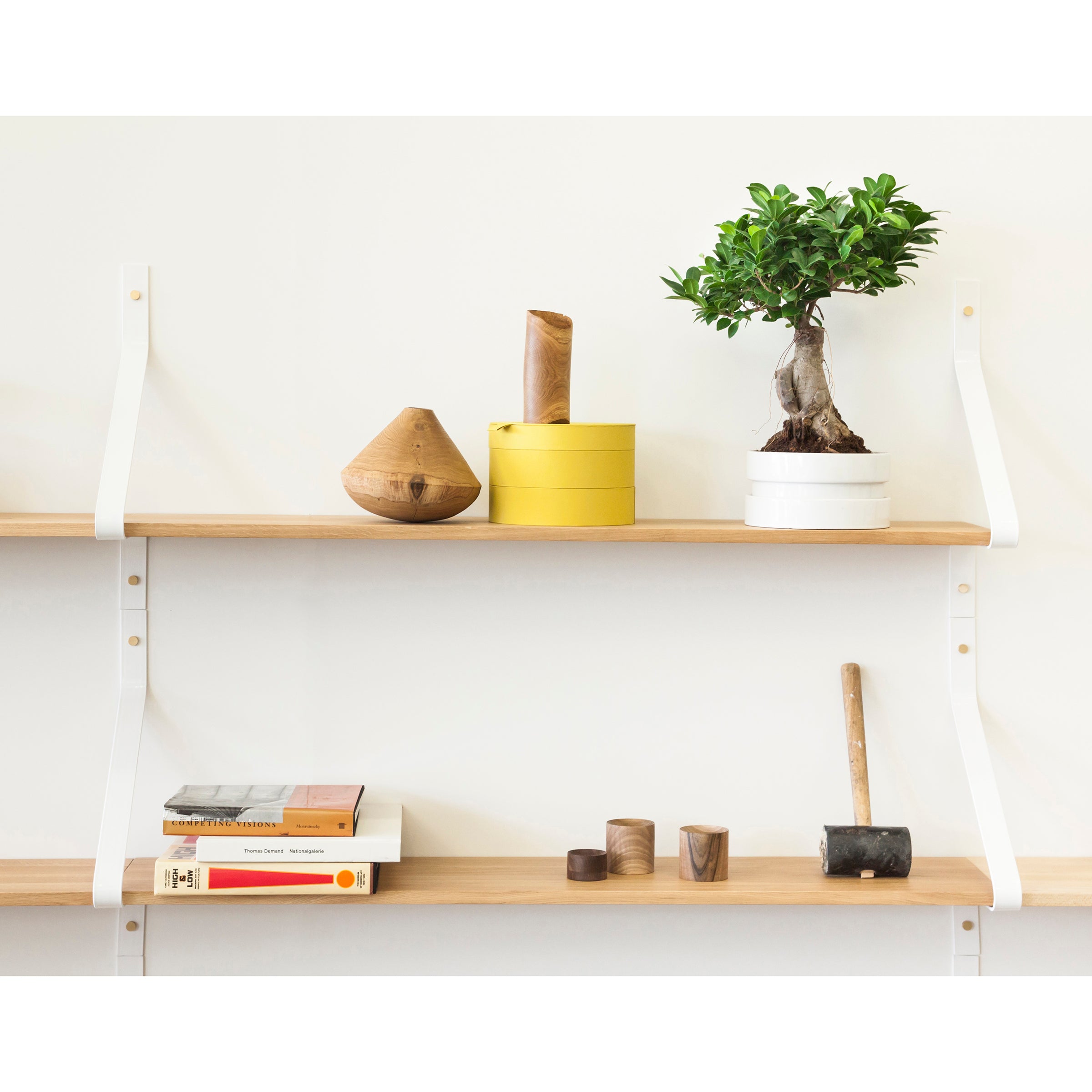 The Ribbon Shelf by One For Hundred features two solid wood shelves with powder-coated steel brackets, showcasing a small tree in a round pot, cylindrical containers, wooden ornaments, books, and a mallet on the lower shelf against a white wall.