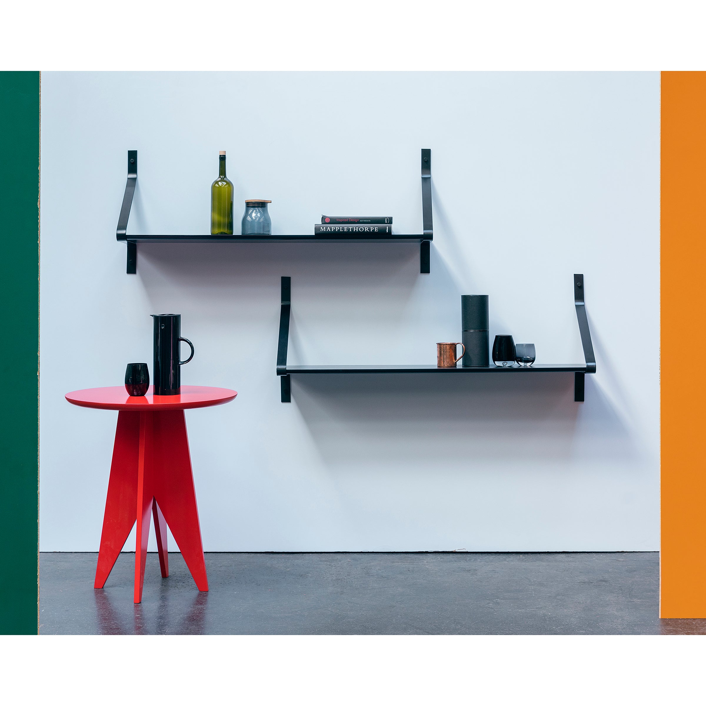The minimalist interior showcases One For Hundreds Ribbon Shelf with powder-coated steel brackets, holding a wine bottle, books, and decor above a small red round table with black cups. The white wall is accented in green and orange hues, complemented by the gray-finished floor.