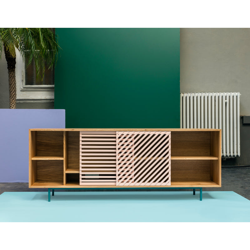 The Shadow Sideboard by One For Hundred is elegantly crafted from solid wood and features open shelves, two decorative pink slatted sliding doors, and minimalist metal legs against a geometric backdrop of green, blue, and purple panels beside a white radiator and palm plant.