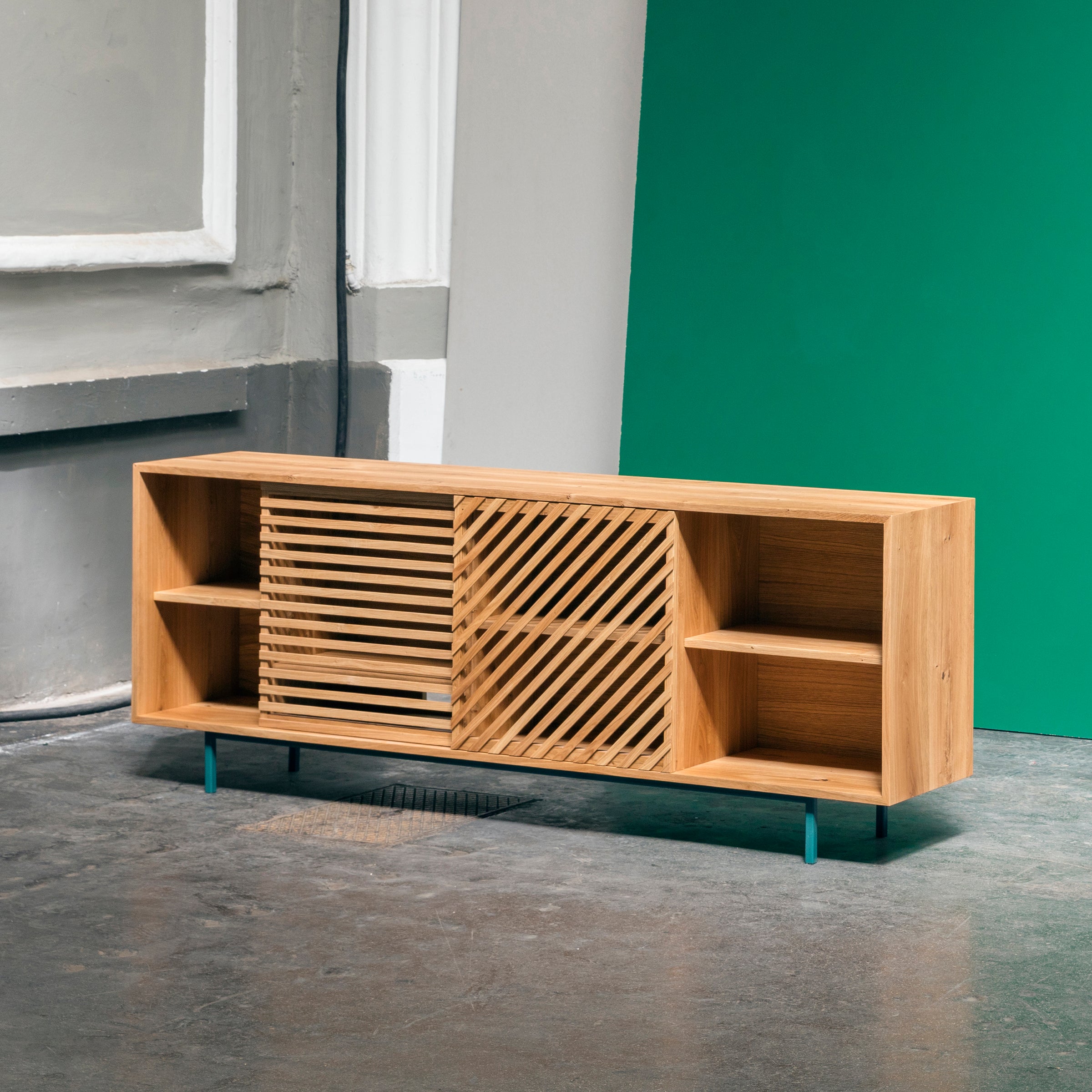 The One For Hundred Shadow Sideboard, a modern piece featuring an open design and two central sliding slatted doors, stands on thin teal legs against gray walls and a green backdrop, with its solid wood elegance enhanced by the polished concrete floor.