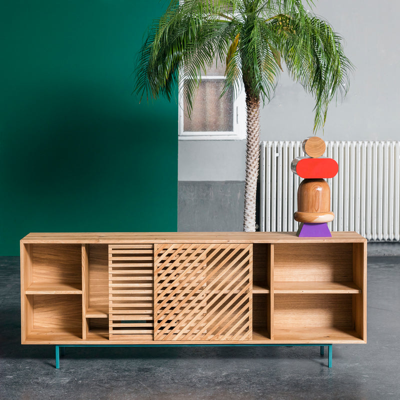 The One For Hundred Shadow Sideboard, crafted from solid wood with open shelves and decorative sliding doors, stands on a gray floor. A geometric sculpture is on top. In the background are a green wall, window, radiator, and potted palm tree.