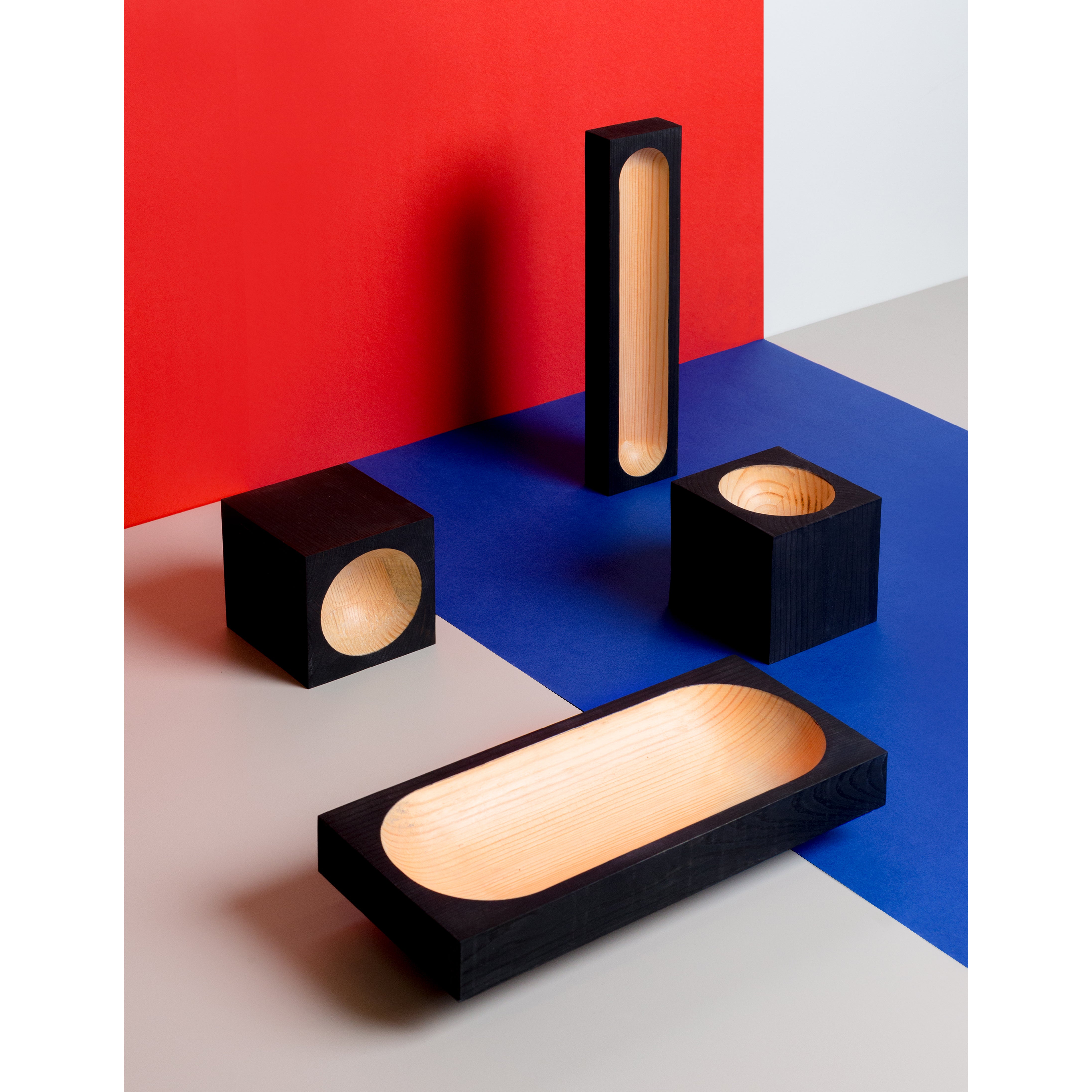 Cavetto by One For Hundred features black pinewood geometric sculptures with hollow centers on a multicolored surface of red, blue, and white. The collection includes a tall rectangle, a square, and elongated ovals, epitomizing minimalist design suitable for table accessories.