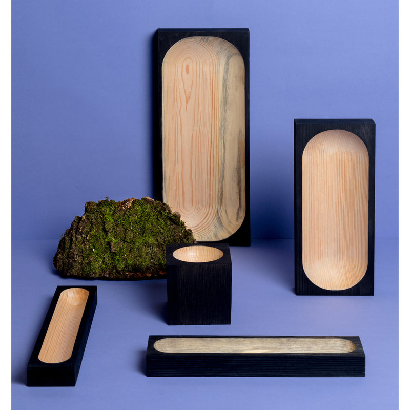 The Cavetto collection by One For Hundred showcases black wooden trays with natural pinewood interiors in various shapes and sizes on a purple background, complemented by artfully placed moss-covered bark.