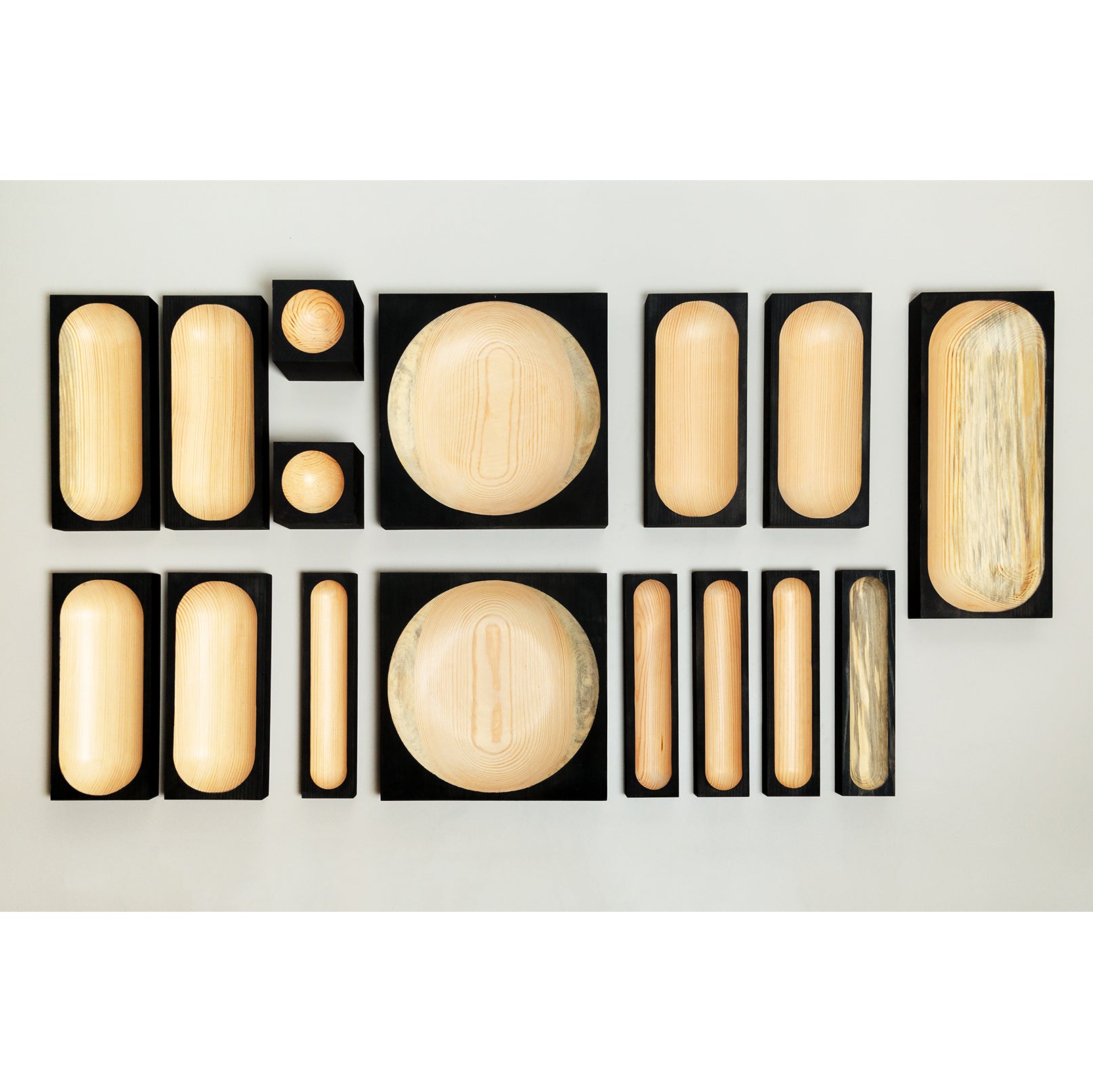 The Cavetto by One For Hundred showcases assorted smooth, oval-shaped pinewood items. Each is set in a cavetto-edged black frame on a gray background, varying from round to elongated ovals, and serve as elegant table accessories.
