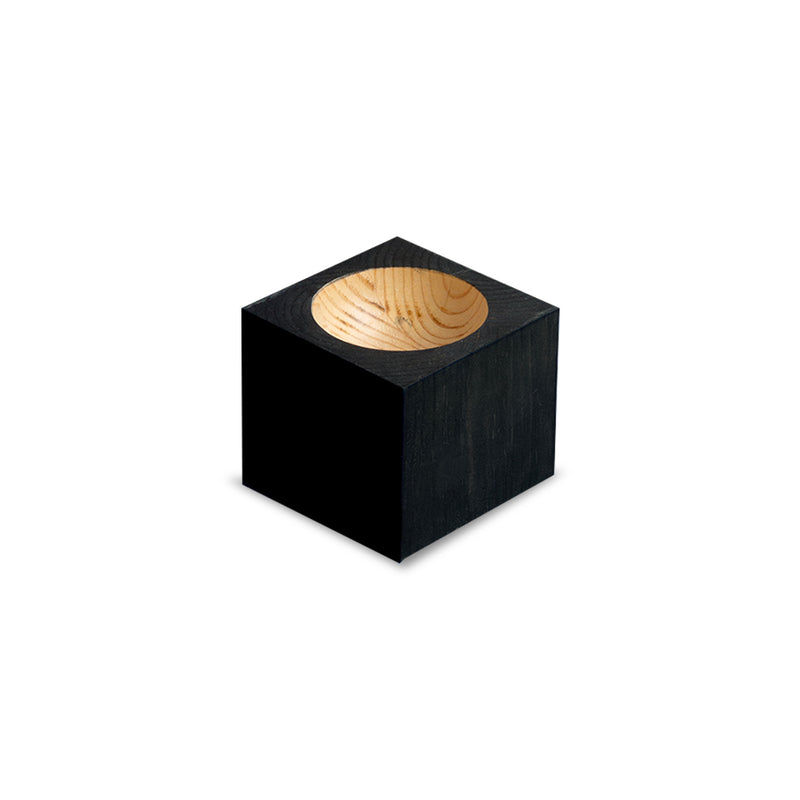 The Cavetto by One For Hundred is a small cubic wooden block made of rich pinewood, with a smooth rounded cavetto top that reveals its natural grain. The black-painted exterior sharply contrasts with the light wood inside—a sophisticated table accessory addition.