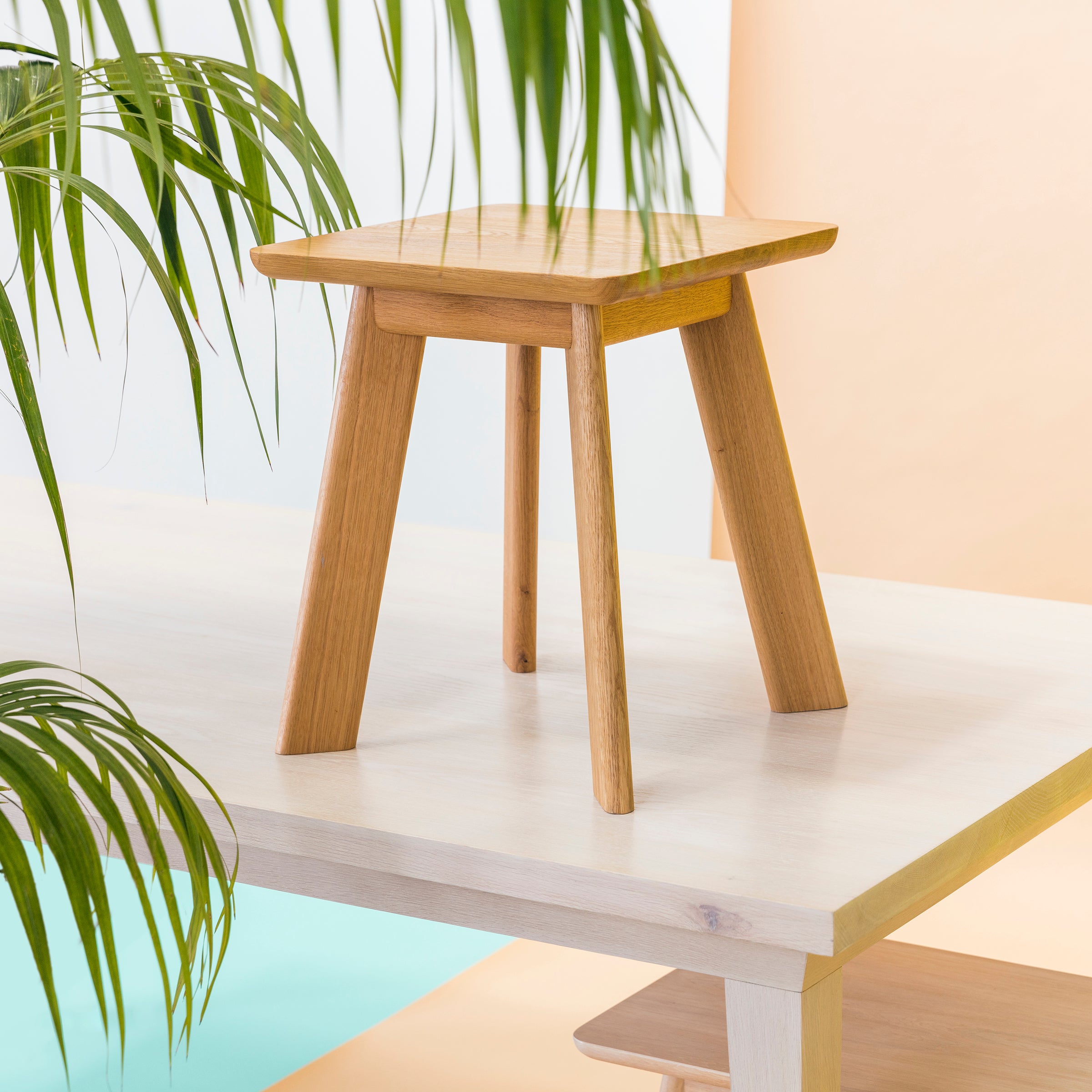 The YOGI Stool by One For Hundred, crafted from solid wood with four angled legs, is displayed on a white table. The scene is framed by green palm leaves against a soft backdrop of pastel light peach and aqua blue.