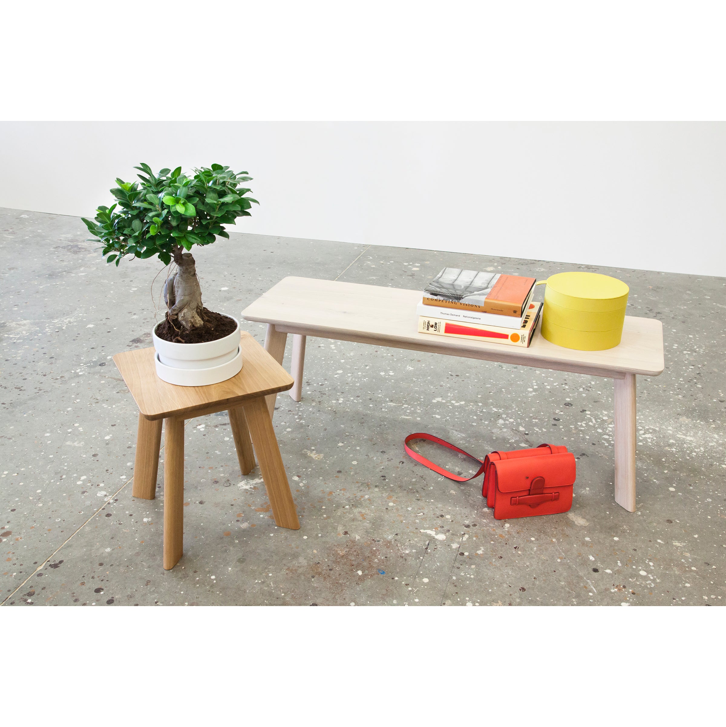 A minimalistic interior scene showcases a small YOGI Stool by One For Hundred, holding a potted plant. A light wooden bench with stacked books and a yellow container complements an orange bag on the concrete floor, highlighting the charm of solid wood furniture.
