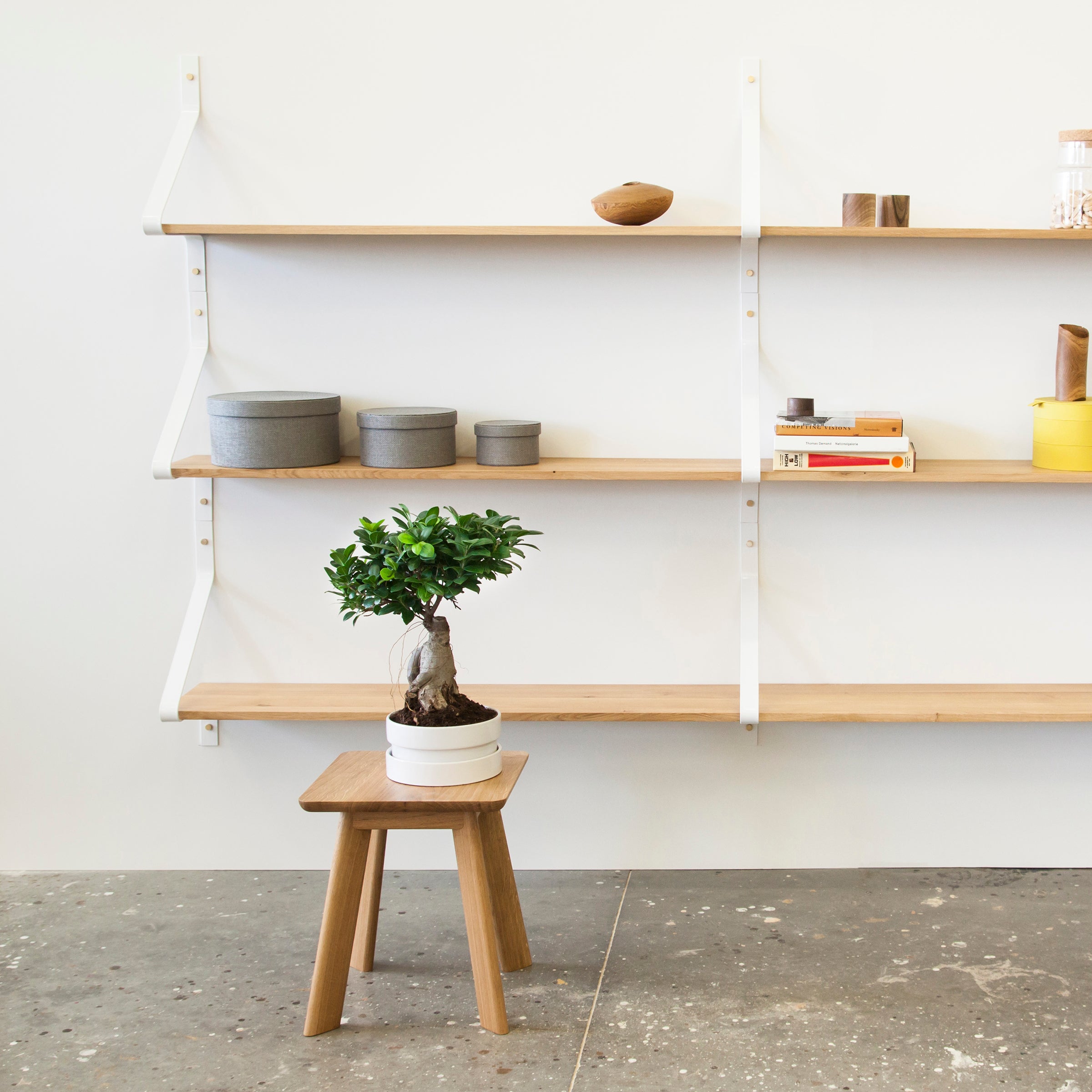 A minimalist interior showcases a small bonsai on a YOGI Stool by One For Hundred. White wall-mounted shelves hold stacked gray boxes, a yellow container, books, and decor items against the backdrop of a concrete floor.