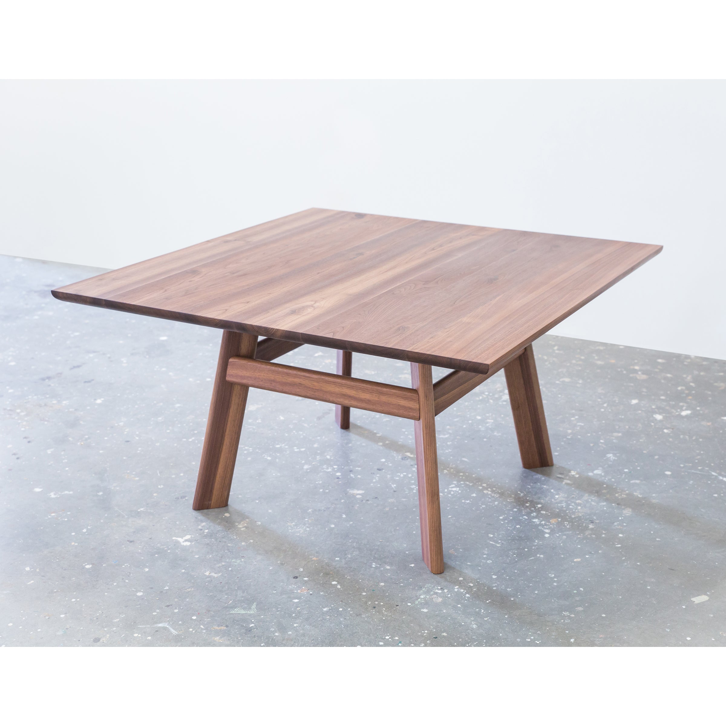 The YOGI Table by One For Hundred is a square wooden piece with a sleek design, smooth surface, and angled legs. It sits elegantly on a polished gray concrete floor against a plain white background.