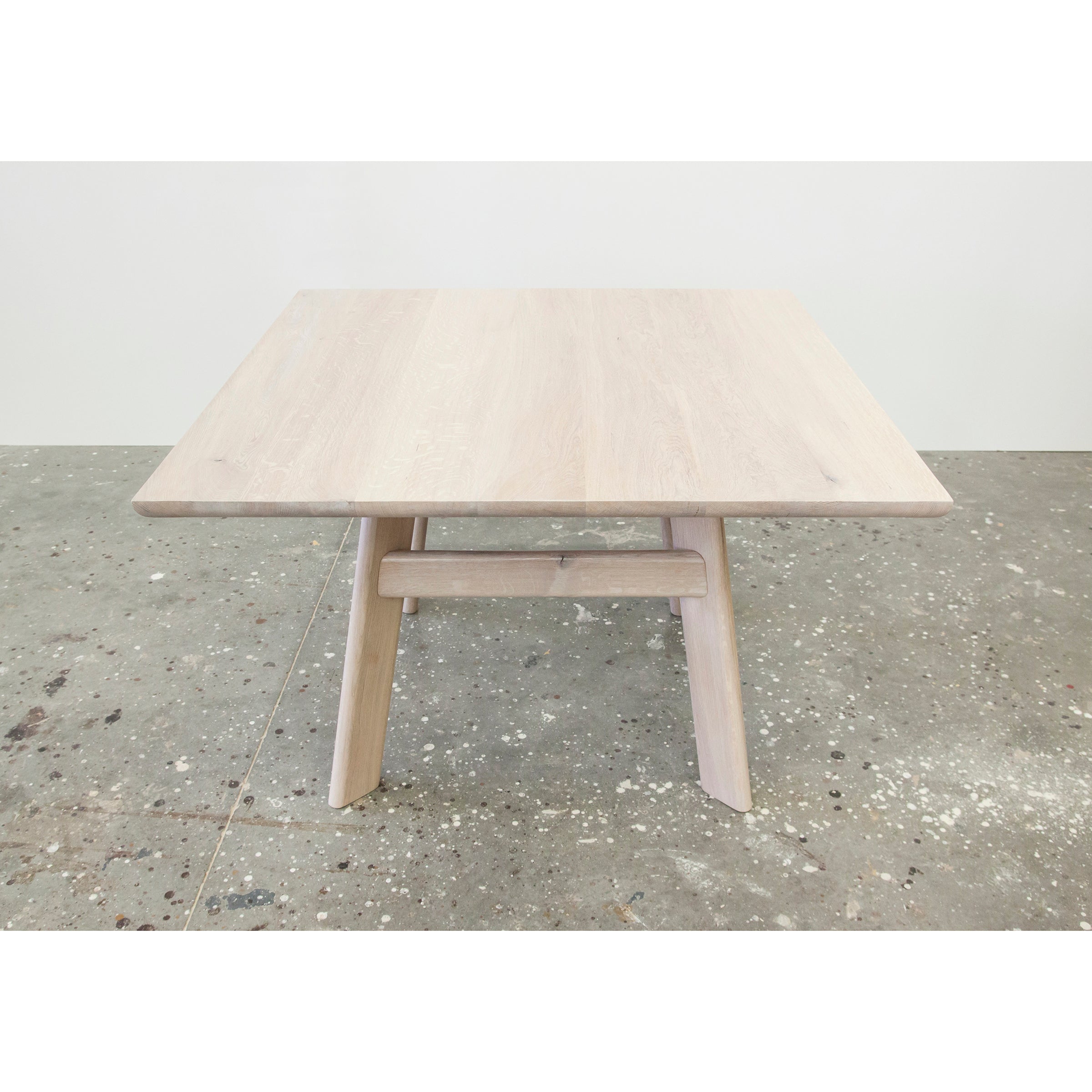A square YOGI Table from One For Hundred, featuring a light wood finish and minimalist design, rests on a speckled concrete floor. It highlights the natural grain with two angled legs and a horizontal support beam for a sturdy, modern base.