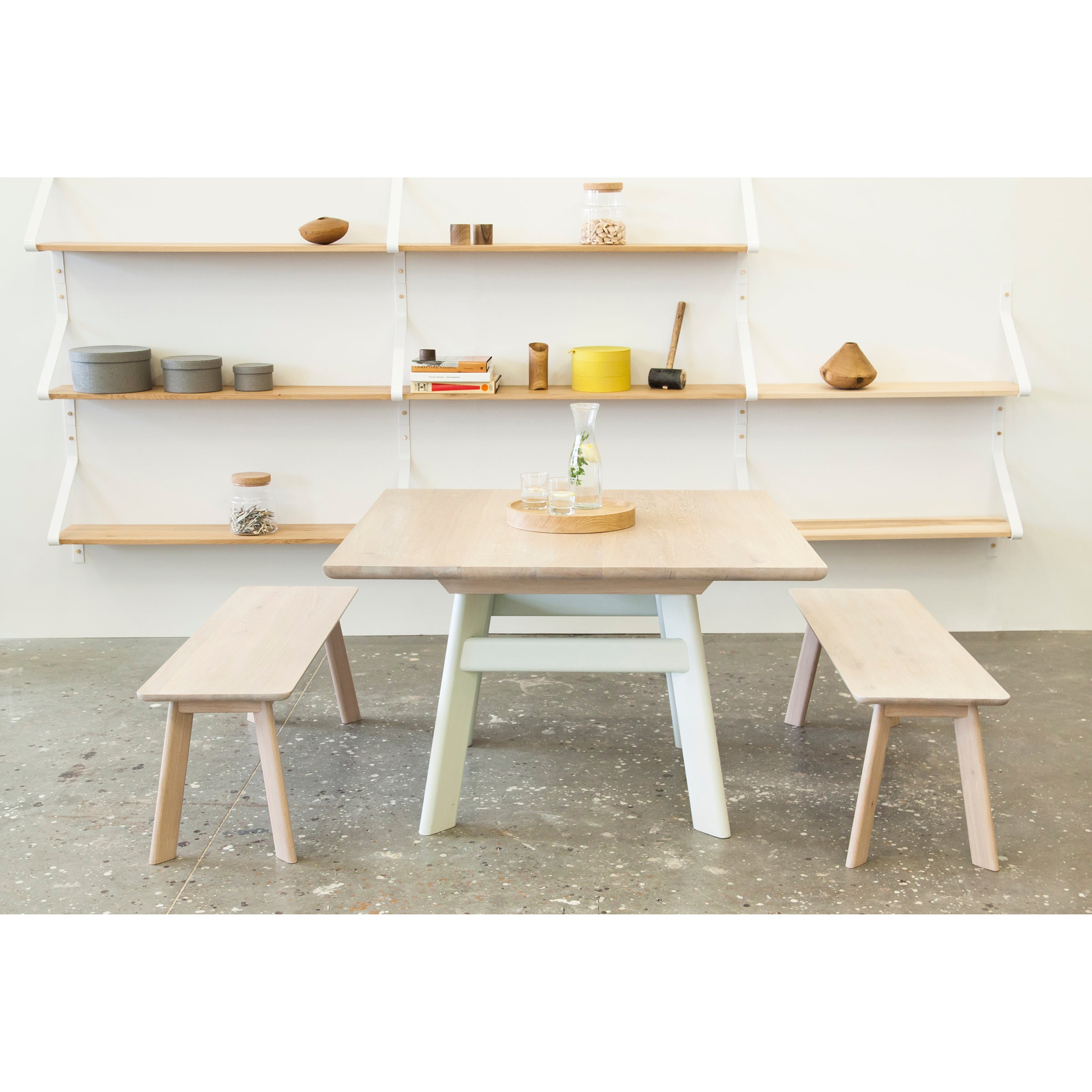A minimalist dining area features a solid wood YOGI Table by One For Hundred, accompanied by two benches. Background shelves display decor items such as bowls, a yellow canister, and books. The concrete floor enhances the modern aesthetic.