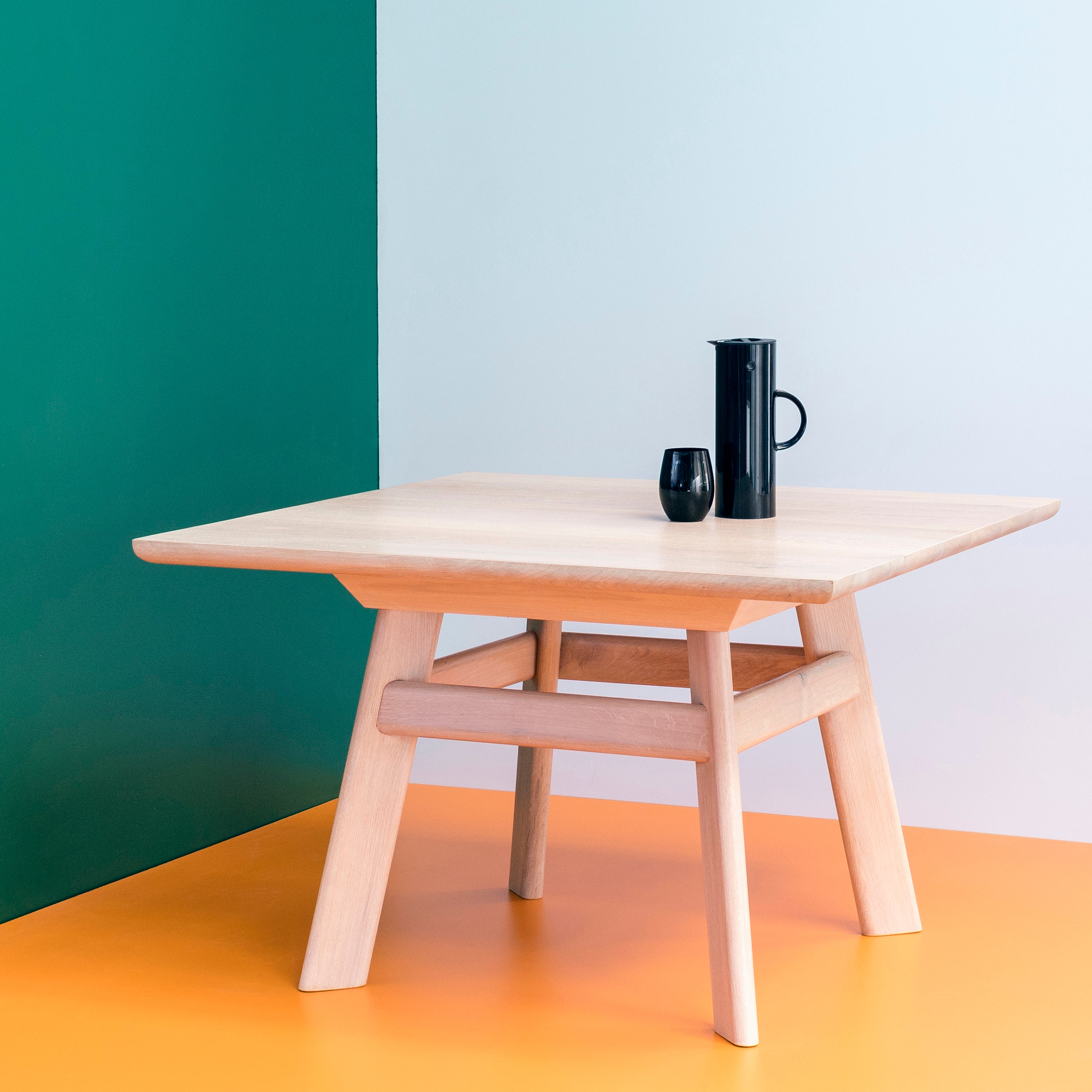 A minimalist solid wood YOGI Table by One For Hundred stands with a light finish on an orange floor, set against a two-toned green and white wall. A black pitcher and a small black cup are elegantly placed on the table.