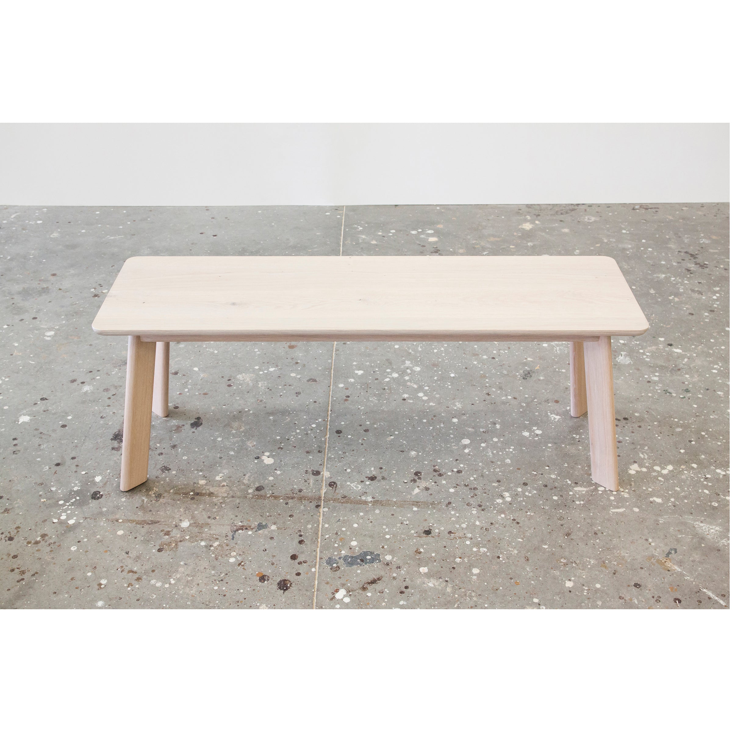 Part of One For Hundreds YOGI collection, this minimalist light wooden YOGI Bench with four legs stands on a speckled concrete floor against a plain white wall.