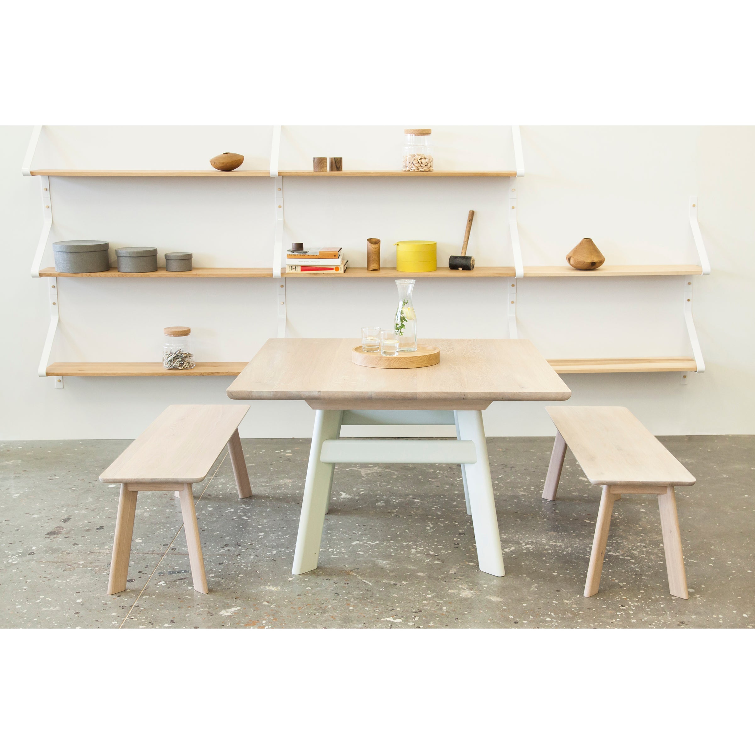 A minimalist dining area showcases a light solid wood table paired with two YOGI Benches from One For Hundred, flanked by a polished concrete floor. In the background, a white wall-mounted shelf displays decorative items, bowls, and jars neatly organized from the YOGI collection.