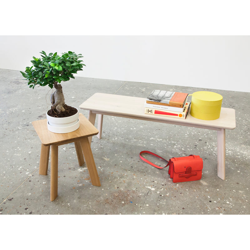A small wooden table from the YOGI collection holds a potted plant. Nearby, the YOGI Bench by One For Hundred, made of solid wood, displays books and a round yellow box. A red shoulder-strap handbag rests on the gray speckled concrete floor.
