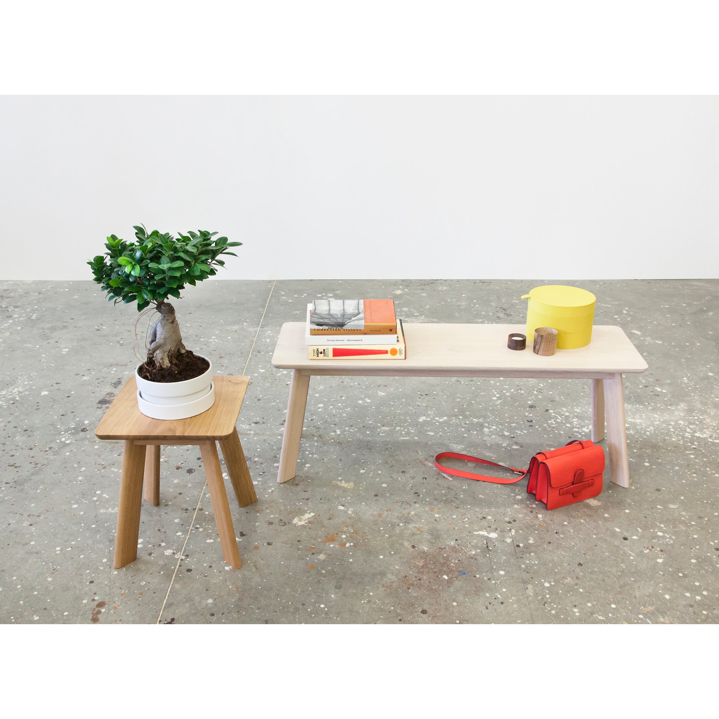 A YOGI Bench by One For Hundred holds books, a yellow container, and a red purse. Next to it, a wooden stool supports a potted bonsai tree. The serene setting features a speckled concrete floor contrasting with the plain white wall.