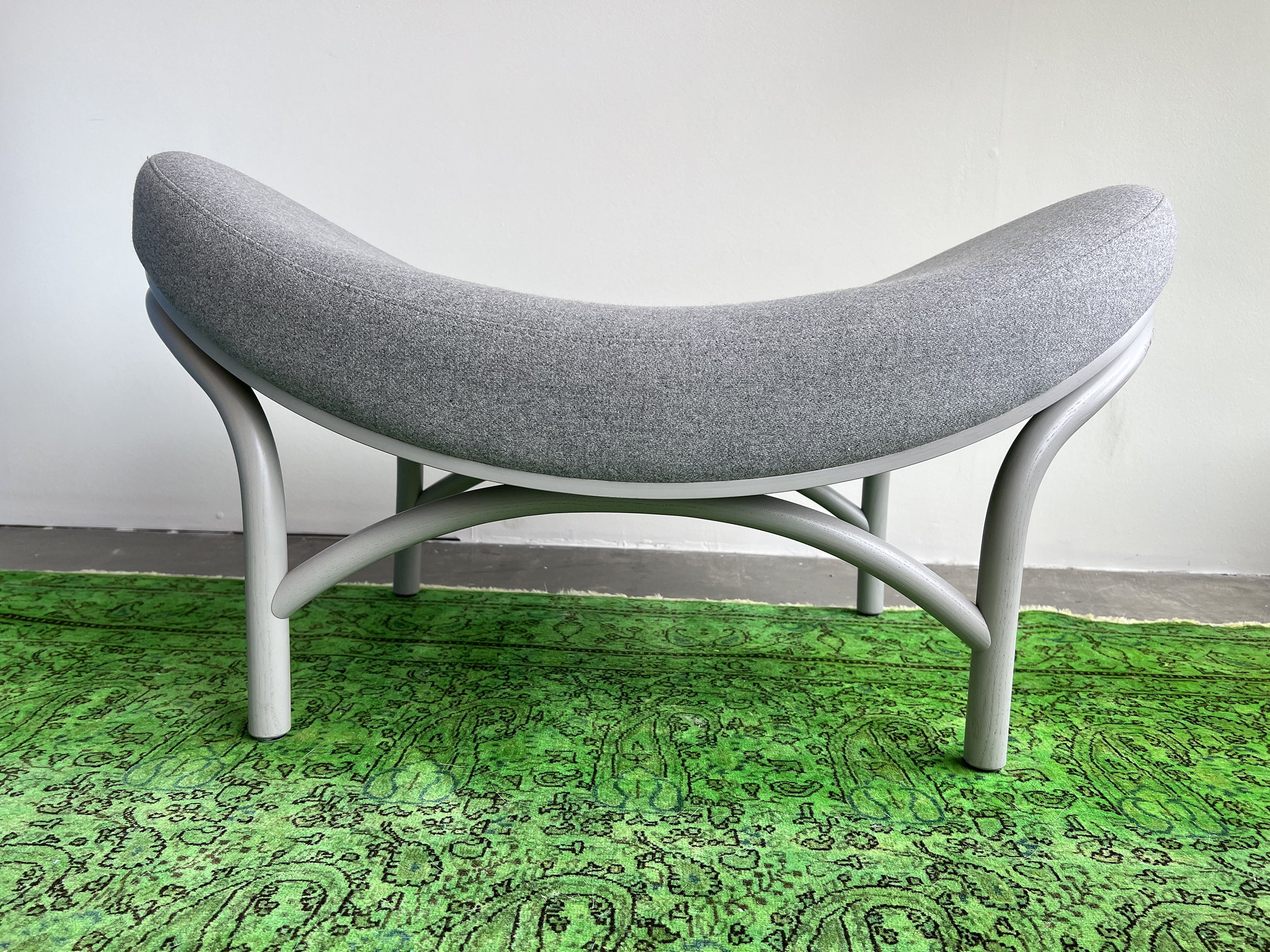 The Ottoman Chips by TON is a modern, crescent-shaped stool featuring a gray upholstered seat on a light gray, manually-bent wood frame. It sits on a vibrant green paisley-patterned rug against a plain white wall.