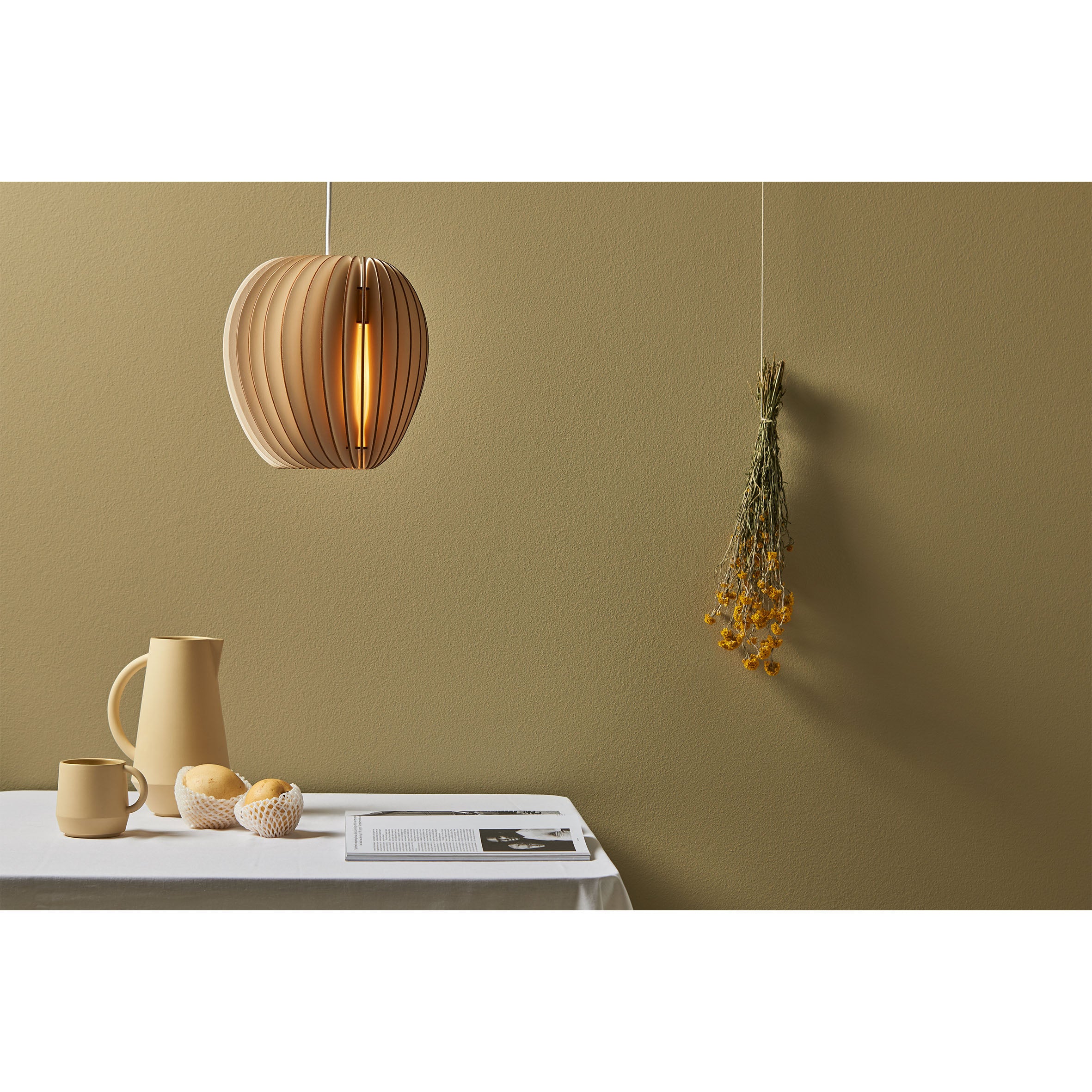 A minimalist dining scene features a beige Schneid Pirum lamp, a white table with cloth, and beige pitcher, mug, bowl of eggs, and book. Yellow flowers on a muted olive-green wall enhance the serene ambiance.