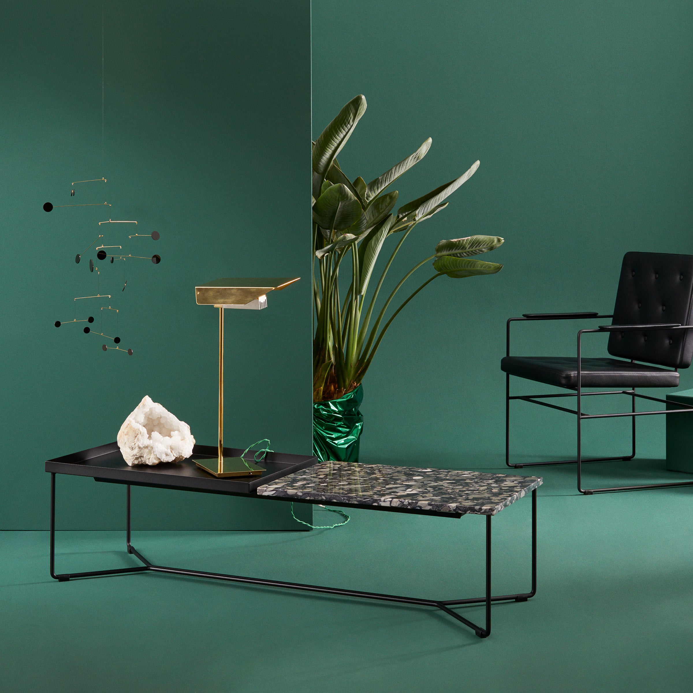 A modern green-themed interior features a black metal chair and a Victor Foxtrot POI Double Marble Coffee Table with Carrara Venatino marble. Nearby, a gold lamp, green-leaf plant in a glass vase, and minimalistic mobile complement the serene green wall and floor.