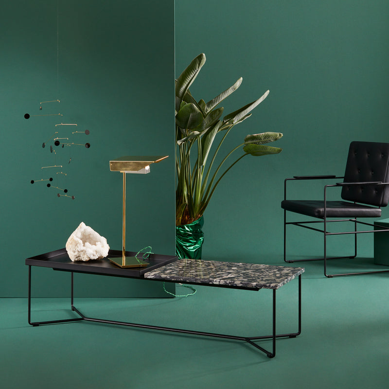 A modern green-themed interior features a black metal chair and a Victor Foxtrot POI Double Marble Coffee Table with Carrara Venatino marble. Nearby, a gold lamp, green-leaf plant in a glass vase, and minimalistic mobile complement the serene green wall and floor.