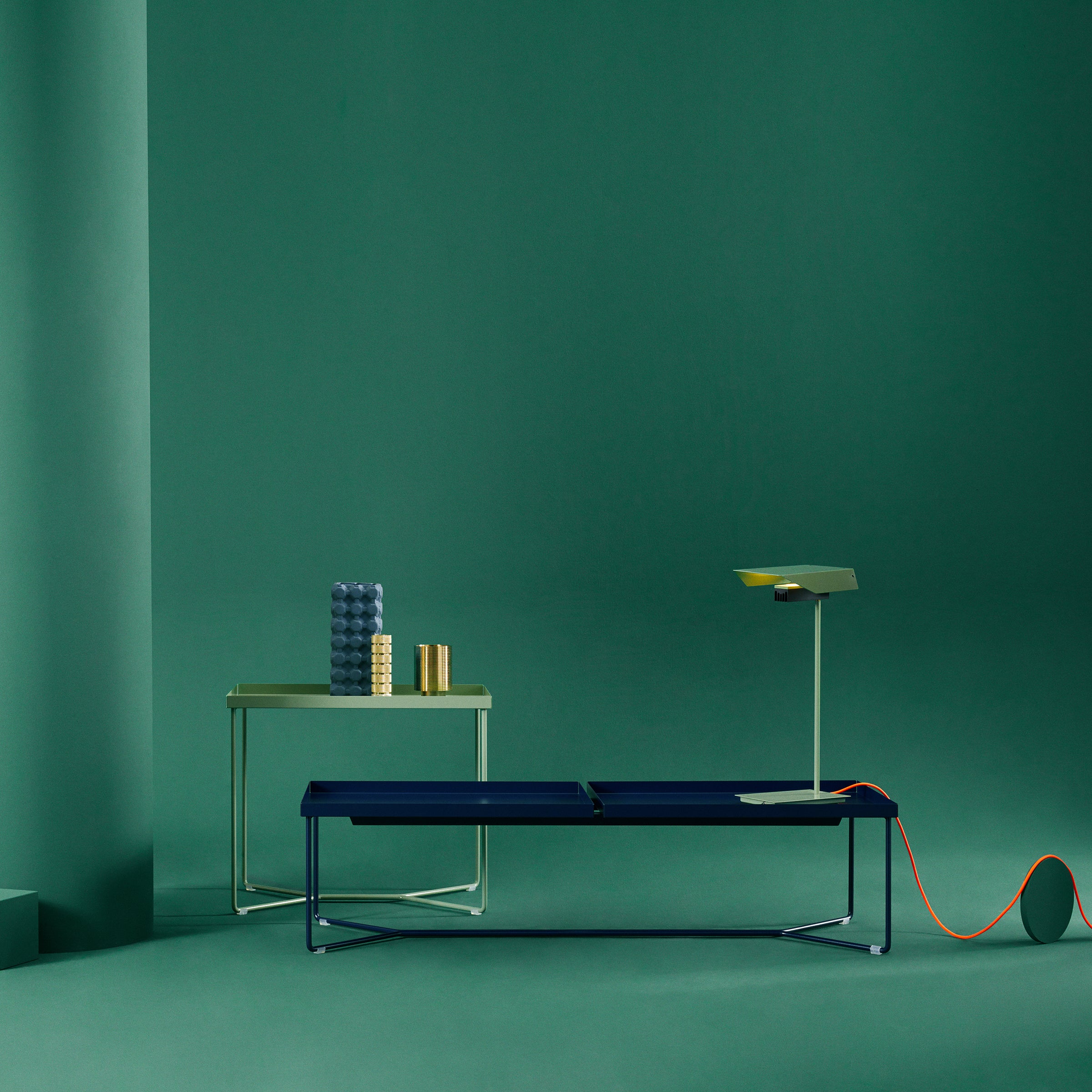 A minimalist interior scene showcases a green backdrop with a sleek Victor Foxtrot POI DOUBLE coffee table displaying round decor, complemented by a modern bench with leather inlays and a stylish lamp featuring an orange cord. The clean, contemporary design is highlighted by steel frame accents.