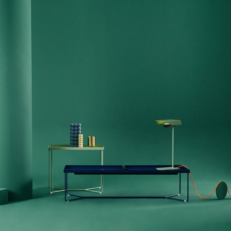 A minimalist interior scene showcases a green backdrop with a sleek Victor Foxtrot POI DOUBLE coffee table displaying round decor, complemented by a modern bench with leather inlays and a stylish lamp featuring an orange cord. The clean, contemporary design is highlighted by steel frame accents.