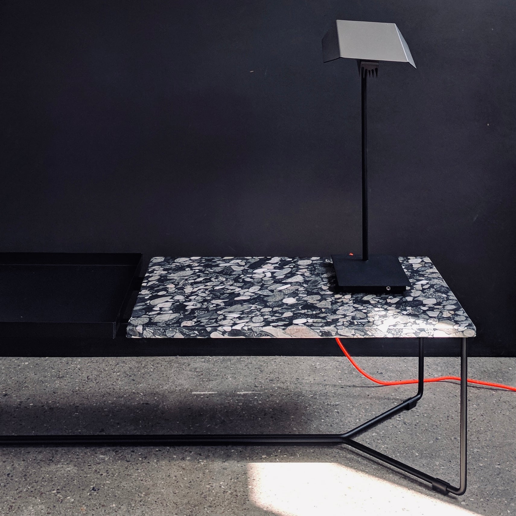 A sleek black lamp sits on the POI DOUBLE MARBLE Coffee Table with Carrara venatino marble by Victor Foxtrot, against a dark wall. A vivid red cable below adds a touch of color.