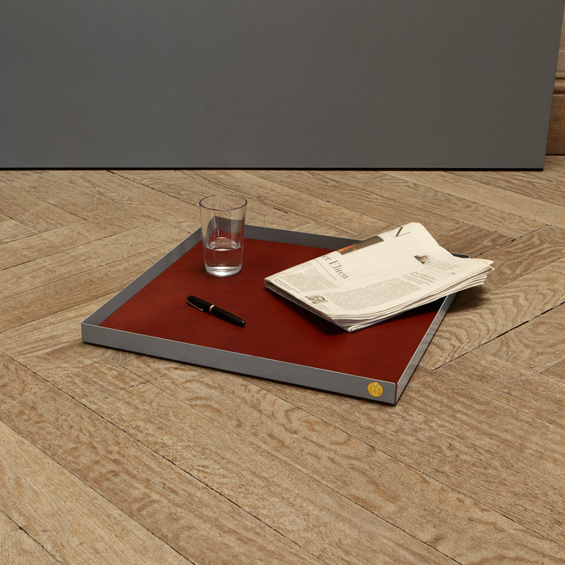 The POI Tray XL by Victor Foxtrot, made from powder-coated steel, sits on a wooden floor. It holds a folded newspaper, a glass of water, and a black pen. A gray wall in the background enhances the minimalist design appeal—ideal for design enthusiasts.