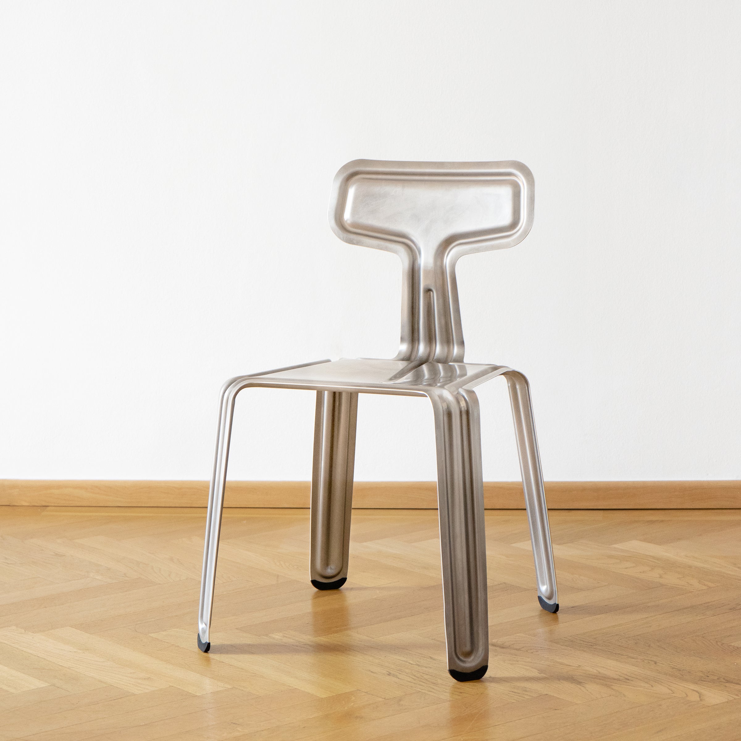 The Moormann Pressed Chair, featuring a modern metallic look with a unique geometric design, stands on a wooden floor. Its T-shaped backrest and thin, flat legs create a futuristic vibe against the plain, light-colored wall. This stackable chair adds style to any space.