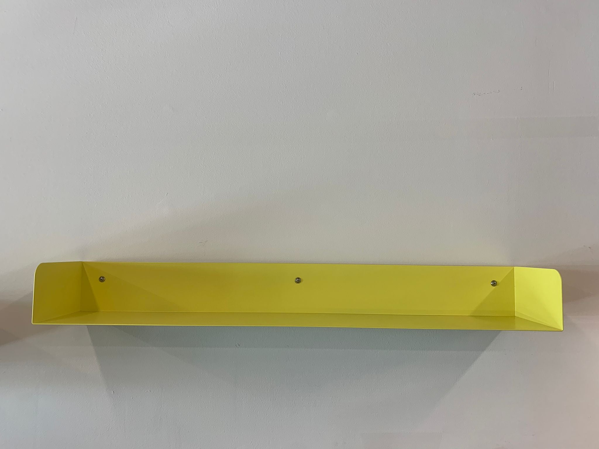 A minimalist Poggibonsi shelf from Atelier Haussmann, in sulfur yellow, is mounted on a white wall with two visible screws. Its smooth surface casts shadows on the wall below.