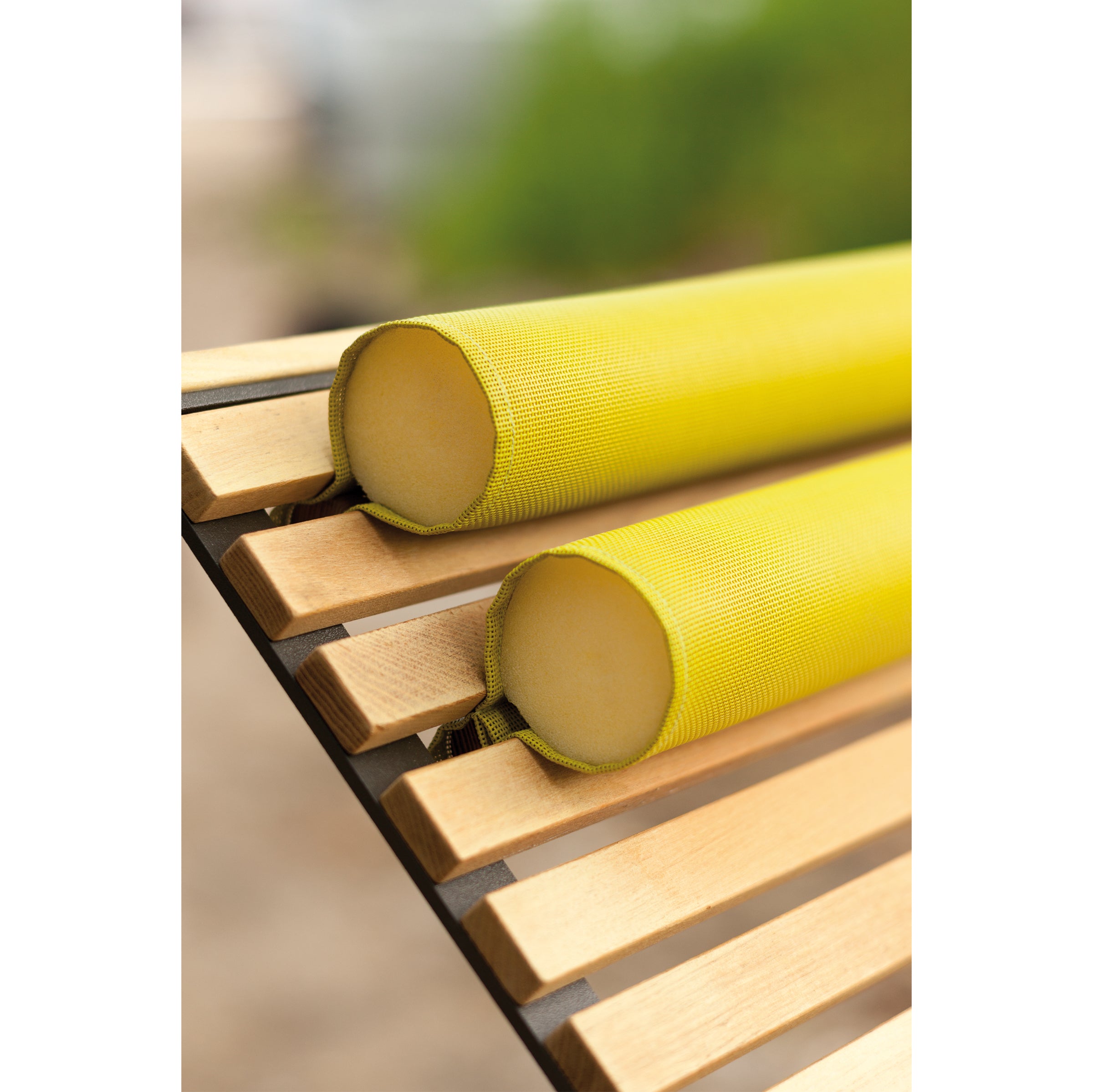 Close-up of two yellow cylindrical cushions from the Egoe Preva Armchair resting on a wooden slatted surface against a blurred green backdrop. The textured fabric cushions, sophisticated in design, feature a looped handle at one end.