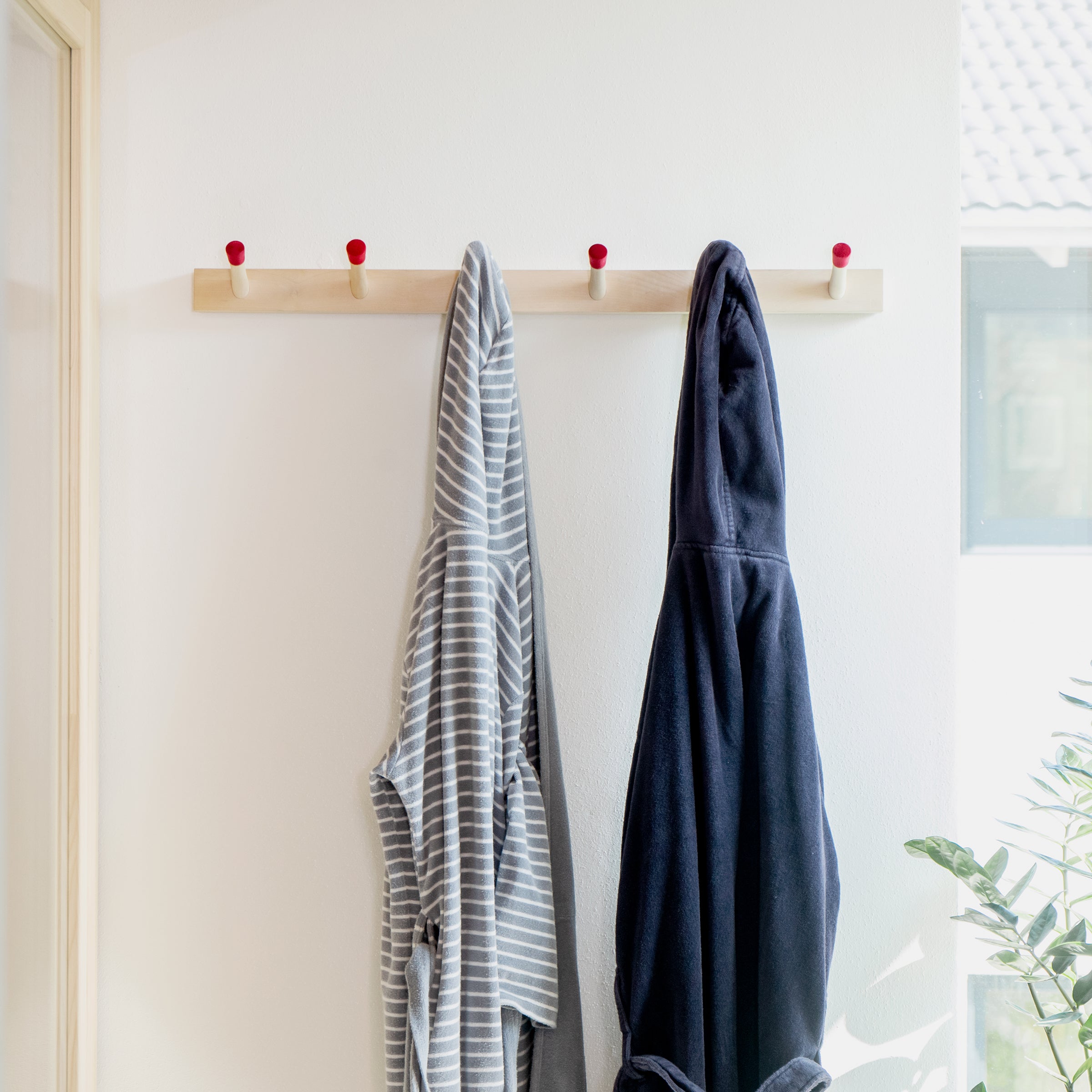 Two Rechenbeispiel bathrobes by Moormann hang on a wooden hook rail against a white wall: one with gray and white stripes, the other solid dark blue. A plant peeks in from the bottom right corner as a window softly illuminates the flexible storage setup in the background.