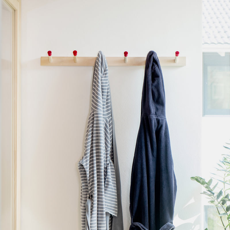 Two Rechenbeispiel bathrobes by Moormann hang on a wooden hook rail against a white wall: one with gray and white stripes, the other solid dark blue. A plant peeks in from the bottom right corner as a window softly illuminates the flexible storage setup in the background.