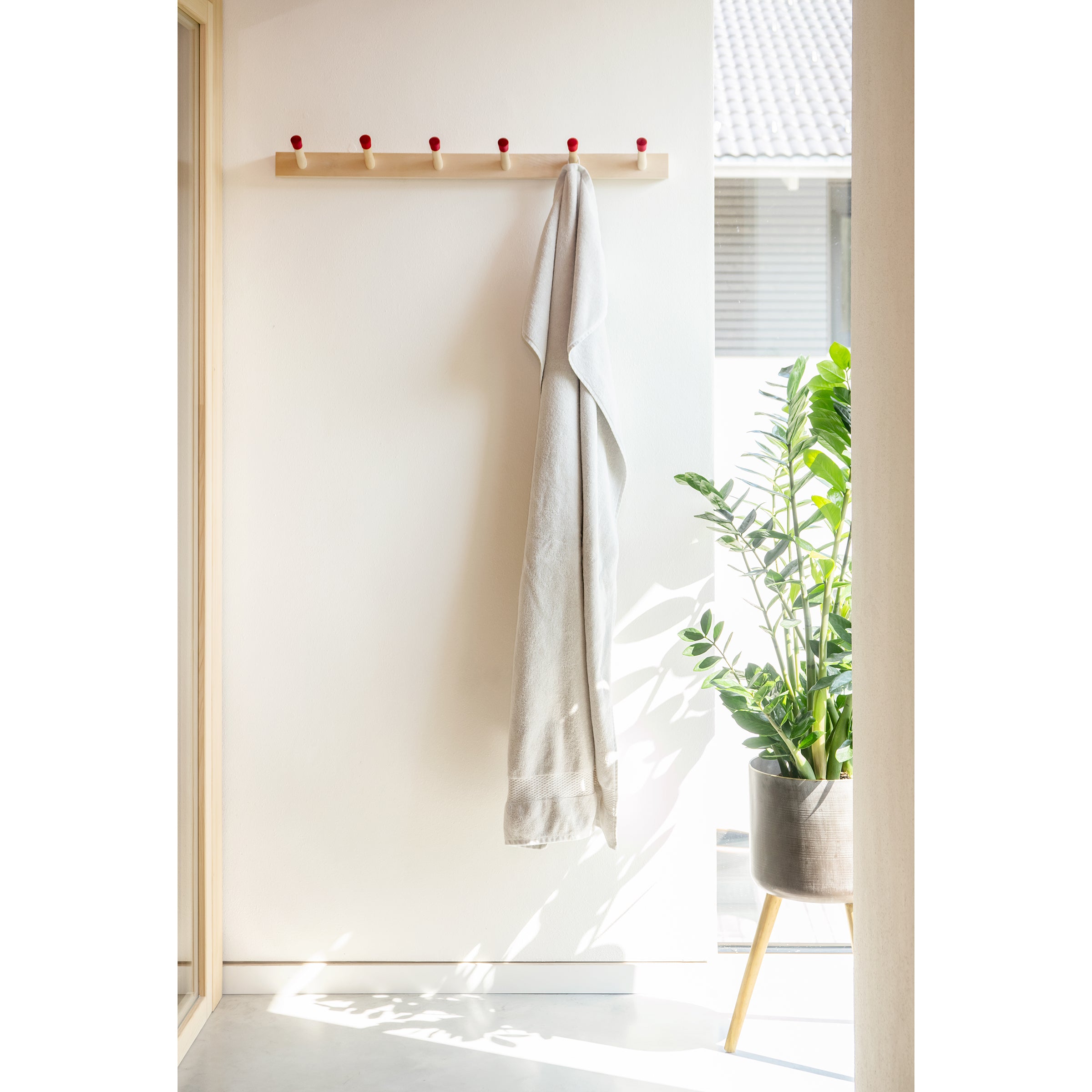 A minimalistic hallway features a Moormann Rechenbeispiel towel rack with light towels on its red pegs against a white wall. Sunlight streams in, casting shadows while a large leafy plant in a modern pot and flexible storage solutions keep the space organized.
