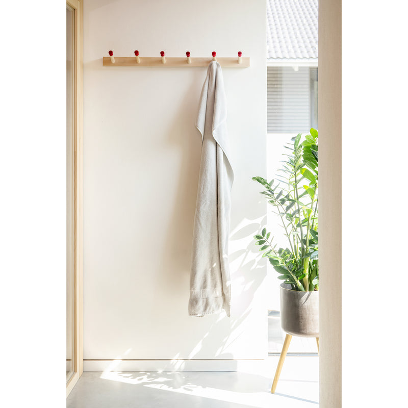 A minimalistic hallway features a Moormann Rechenbeispiel towel rack with light towels on its red pegs against a white wall. Sunlight streams in, casting shadows while a large leafy plant in a modern pot and flexible storage solutions keep the space organized.