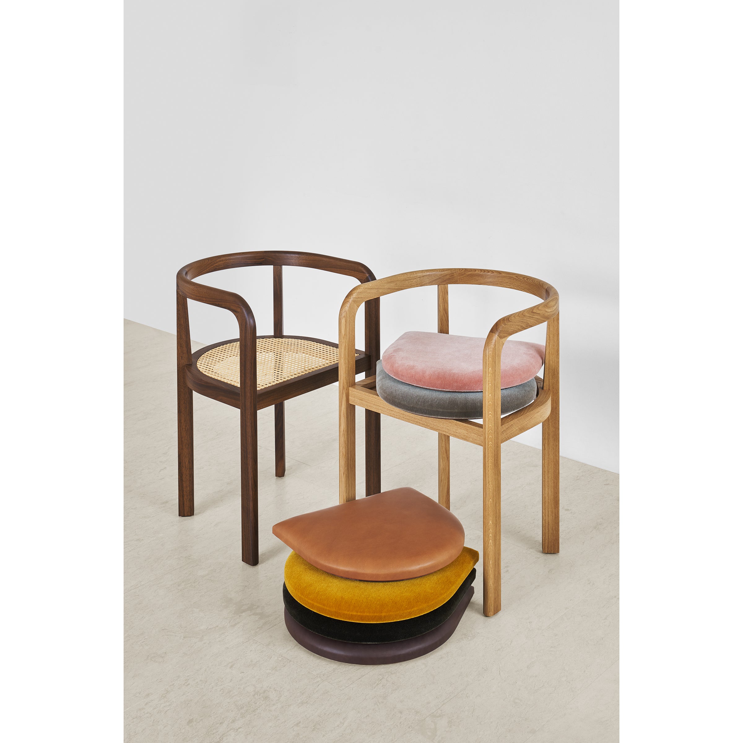 Two RH01 Stuttgart wooden chairs from E15 are showcased. The left chair has a woven seat, and the right features a pink cushion with four colorful teardrop-shaped cushions below. This scene reflects the elegance of Richard Herre design against a plain white wall.