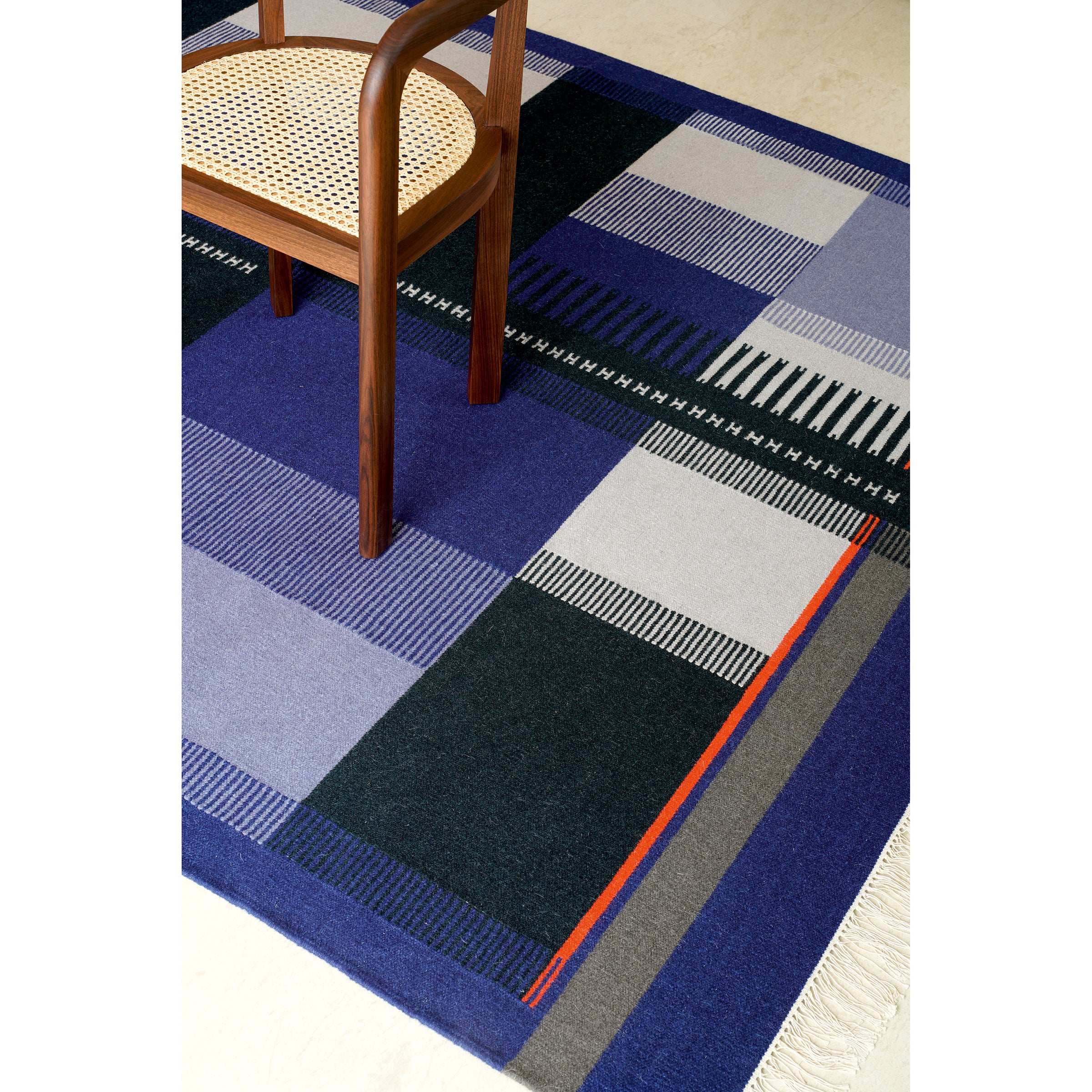 A wooden chair with a woven seat is placed on the RH02 Kilim Zet by E15, made from hand-spun sheep wool. The rug showcases shades of blue, black, and gray with orange accents in a geometric pattern featuring stripes and fringe, reminiscent of Richard Herres artistry.