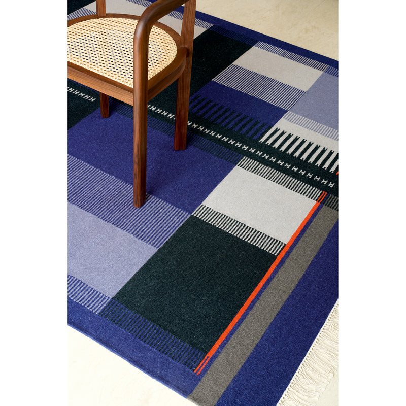 A wooden chair with a woven seat is placed on the RH02 Kilim Zet by E15, made from hand-spun sheep wool. The rug showcases shades of blue, black, and gray with orange accents in a geometric pattern featuring stripes and fringe, reminiscent of Richard Herres artistry.