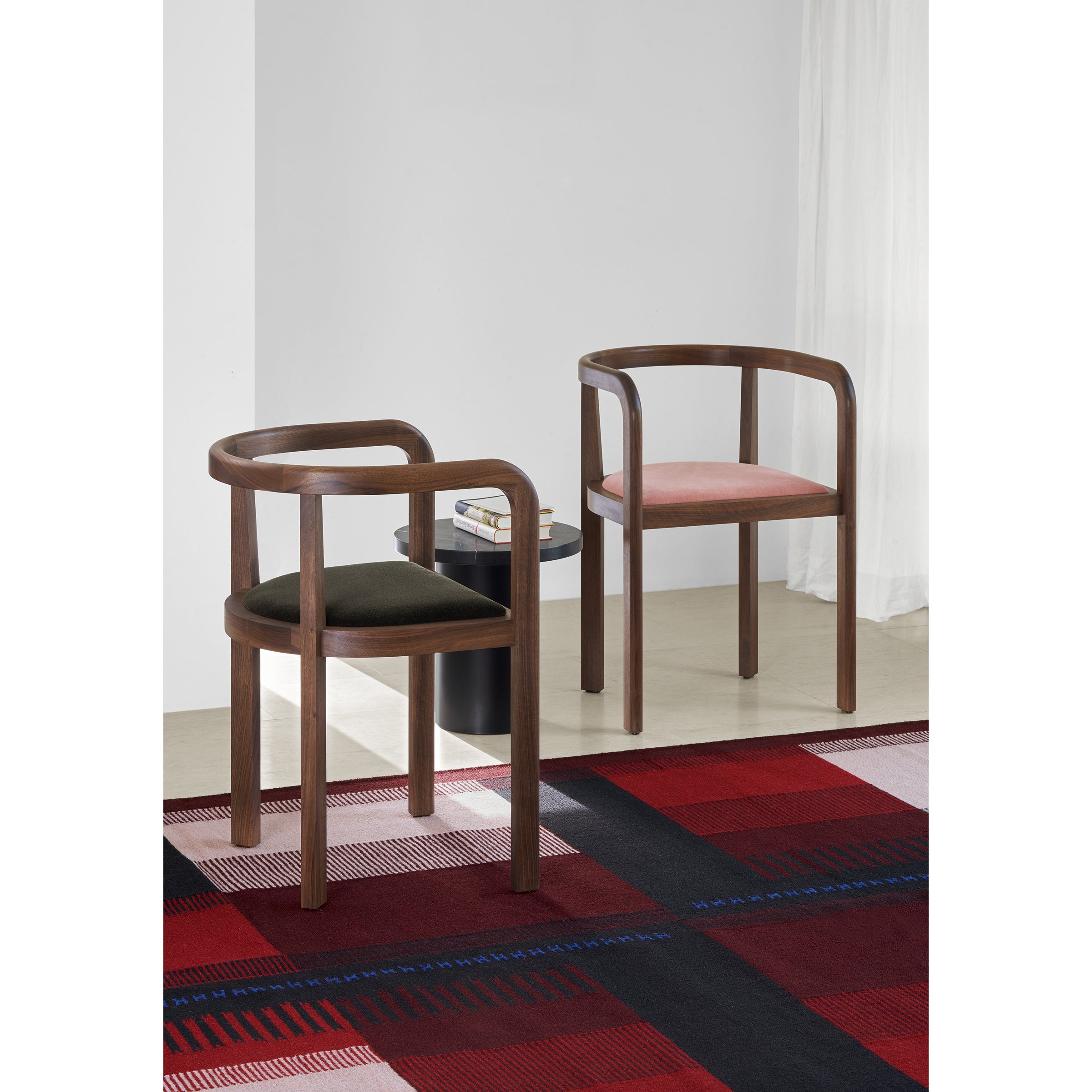 Two E15 RH01 Stuttgart chairs with curved frames and solid wood craftsmanship, one with a dark cushion, the other light, stand beside a small black table holding books. They rest on a red checkered carpet against a sunlit white backdrop, echoing Richard Herres design elegance.