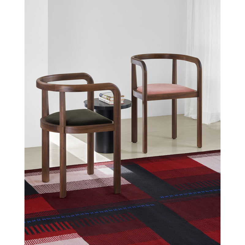 Two wooden chairs with curved armrests and colorful cushions sit beside a small round table holding books. The setting features an RH02 Kilim Zet rug by E15, inspired by Richard Herres red and black checkered design, made from hand-spun sheep wool, with a sheer white curtain in the background.