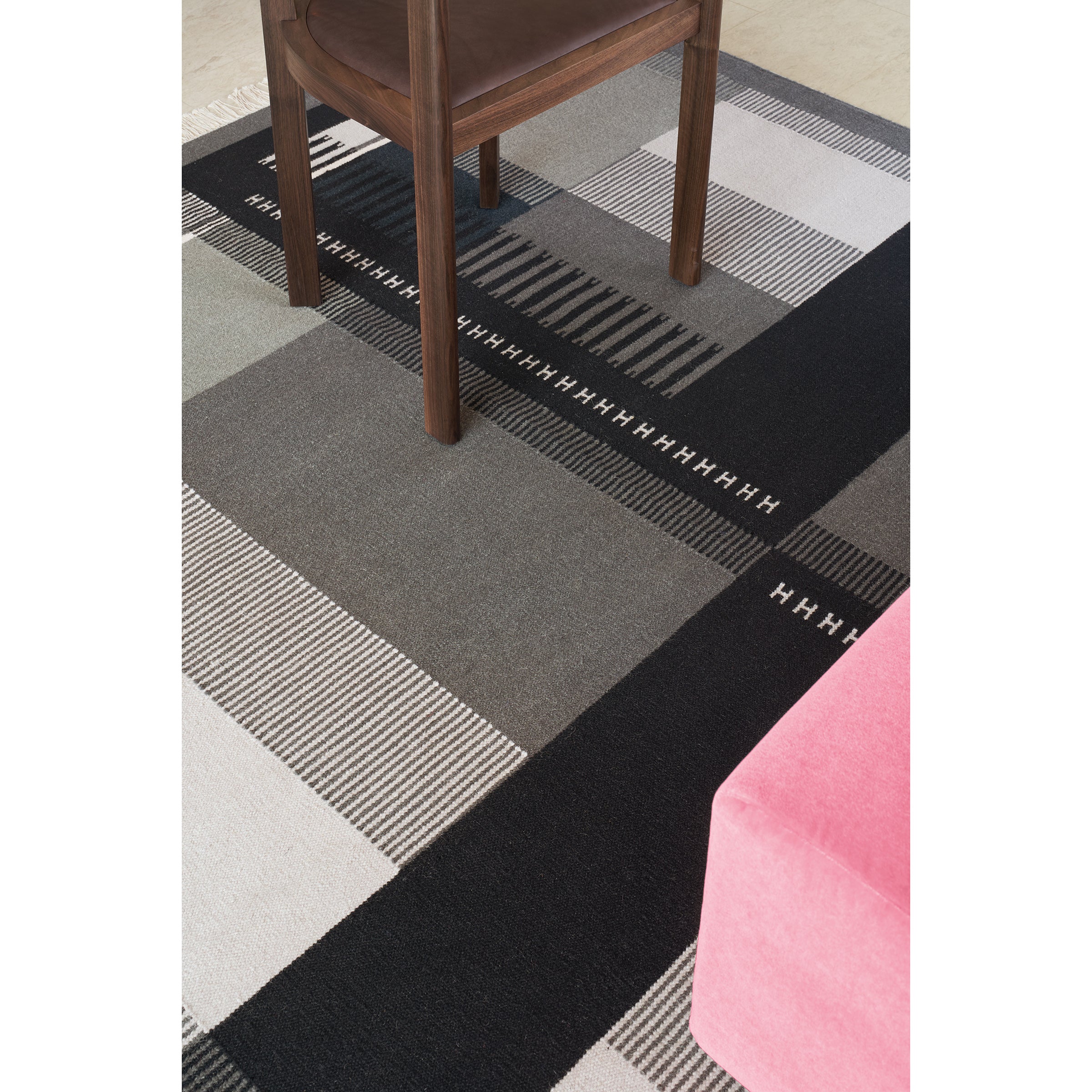 The E15 RH02 Kilim Zet rug, featuring gray, black, and white geometric patterns on a light floor, is crafted from hand-spun sheep wool. It supports a brown wooden chair, with a hint of pink furniture in view.