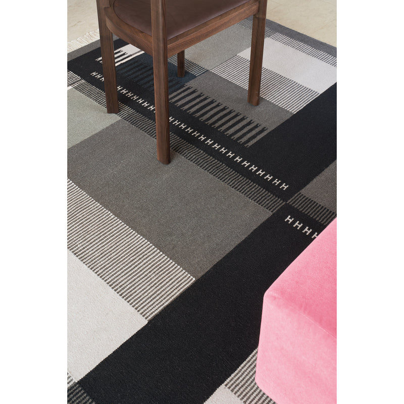 The E15 RH02 Kilim Zet rug, featuring gray, black, and white geometric patterns on a light floor, is crafted from hand-spun sheep wool. It supports a brown wooden chair, with a hint of pink furniture in view.