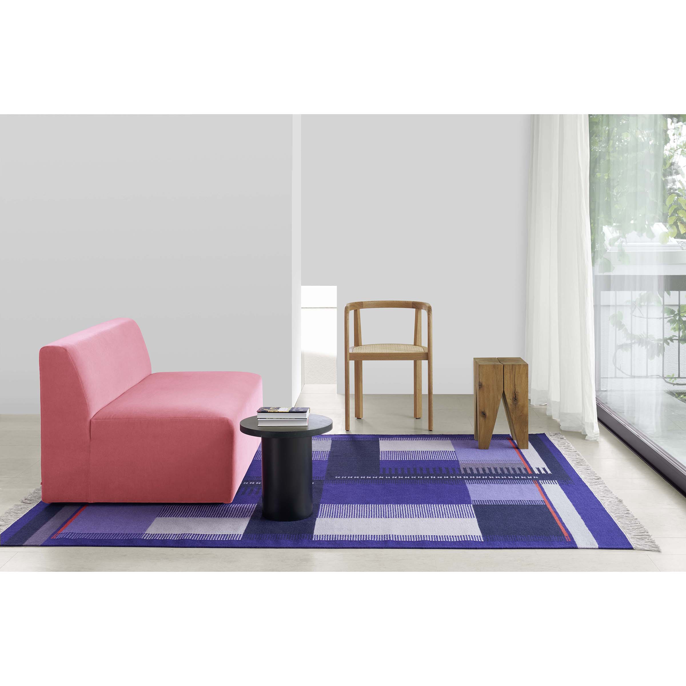 A minimalist space has a pink sofa, wooden chair, small black side table, and unique wooden stool. The floor features an E15 RH02 Kilim Zet blue geometric rug. Sheer curtains on the large window let natural light fill the room.