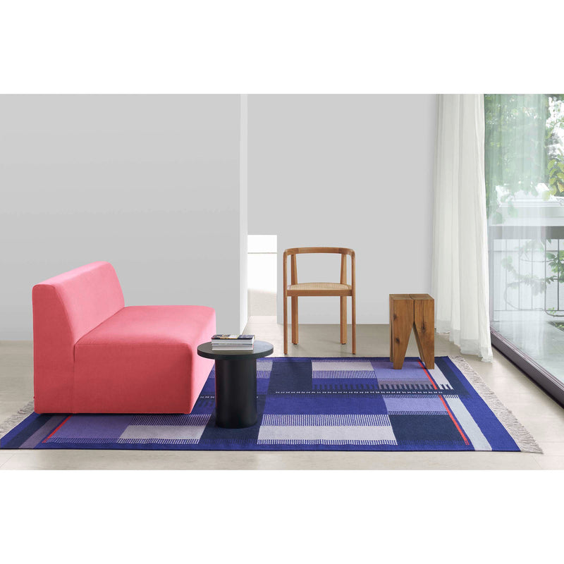A minimalist space has a pink sofa, wooden chair, small black side table, and unique wooden stool. The floor features an E15 RH02 Kilim Zet blue geometric rug. Sheer curtains on the large window let natural light fill the room.