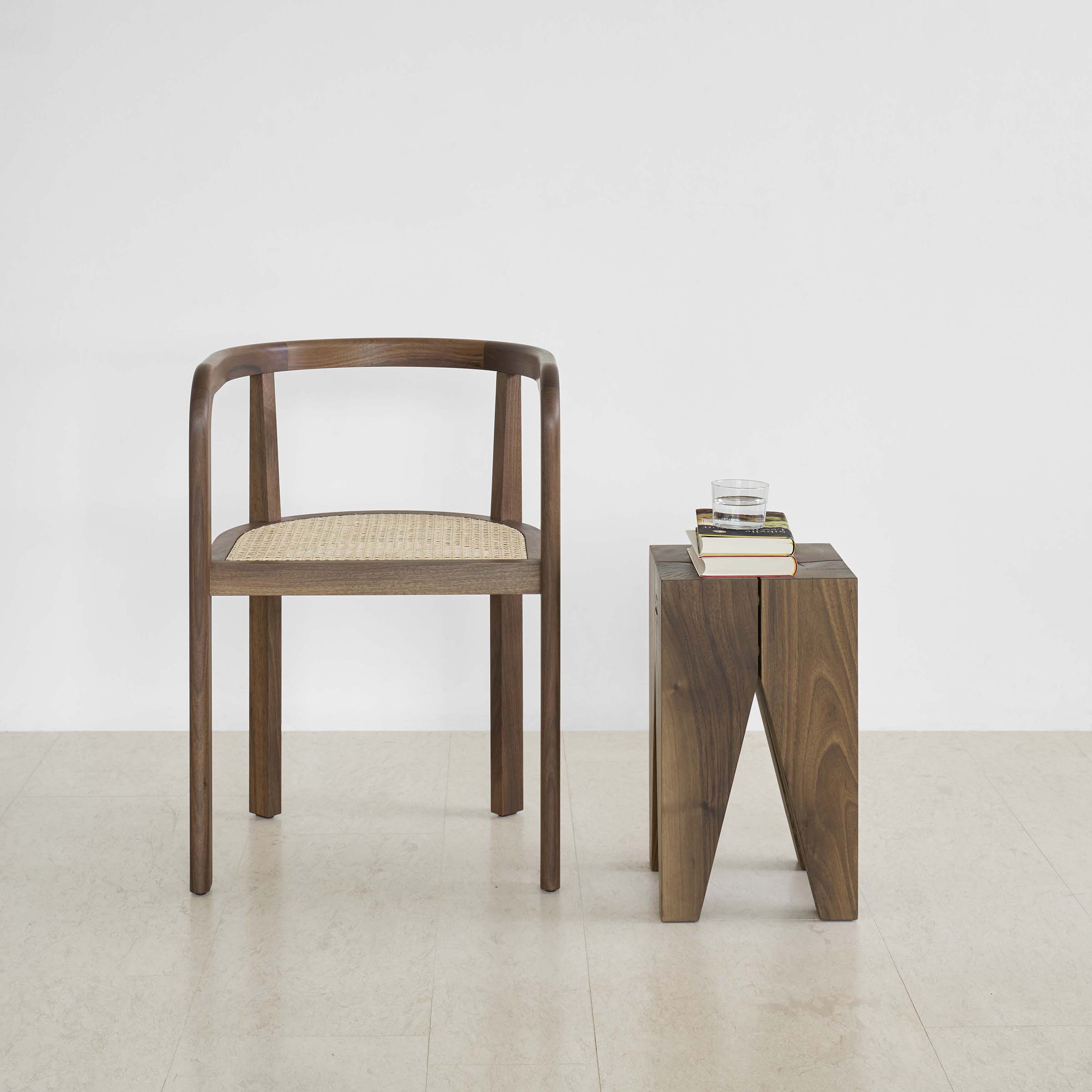 A minimalist interior features an E15 RH01 Stuttgart chair, reflecting classic Richard Herre design with its curved back and woven seat. Next to it, a wooden side table with books and a glass sits against a plain white wall and light-colored floor, exuding solid wood craftsmanship.