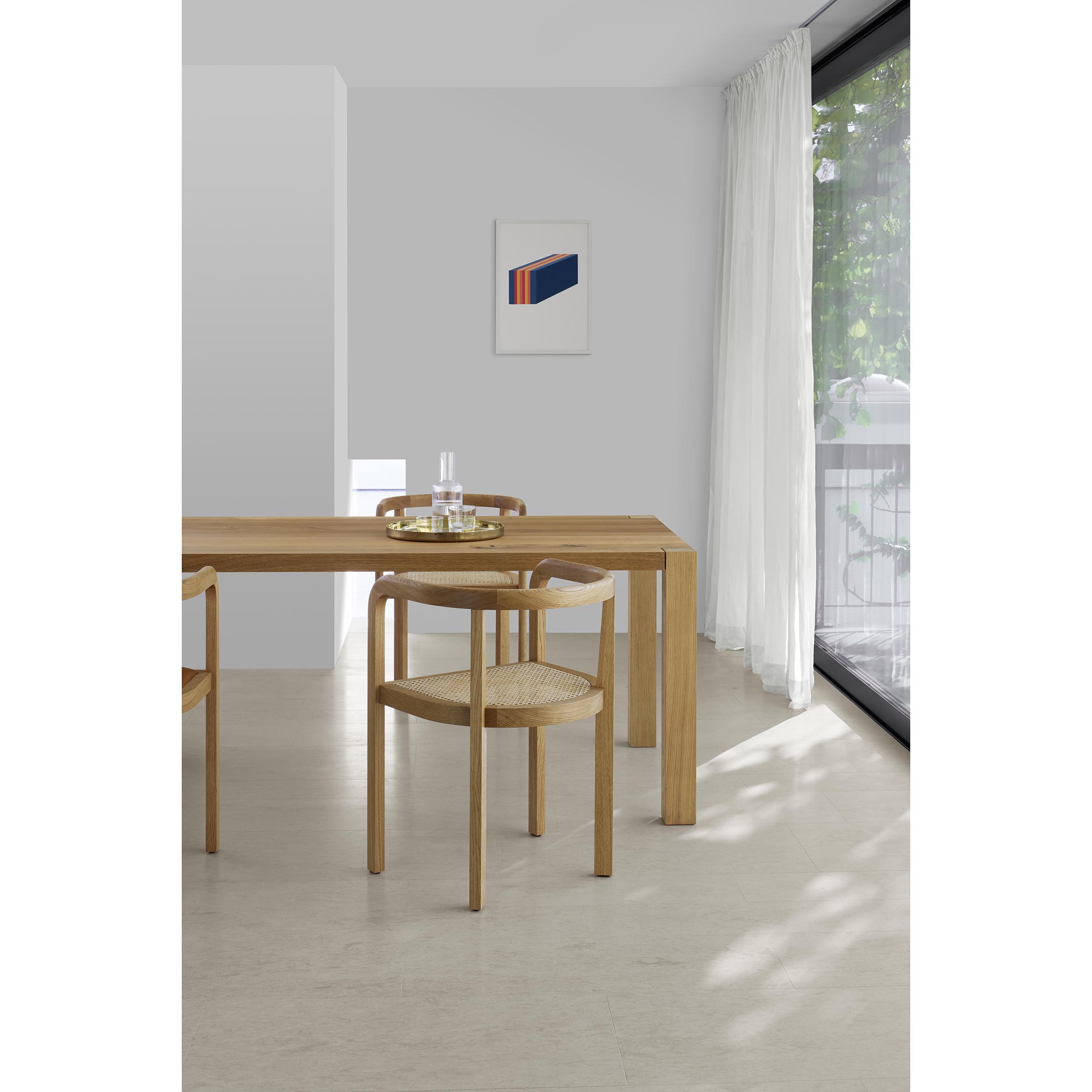 A minimalist dining room with E15s RH01 Stuttgart chairs showcasing Richard Herre design and solid wood craftsmanship, a wooden table, and a glass pitcher set. A colorful abstract painting decorates the light gray wall next to a large window with sheer white curtains overlooking greenery.