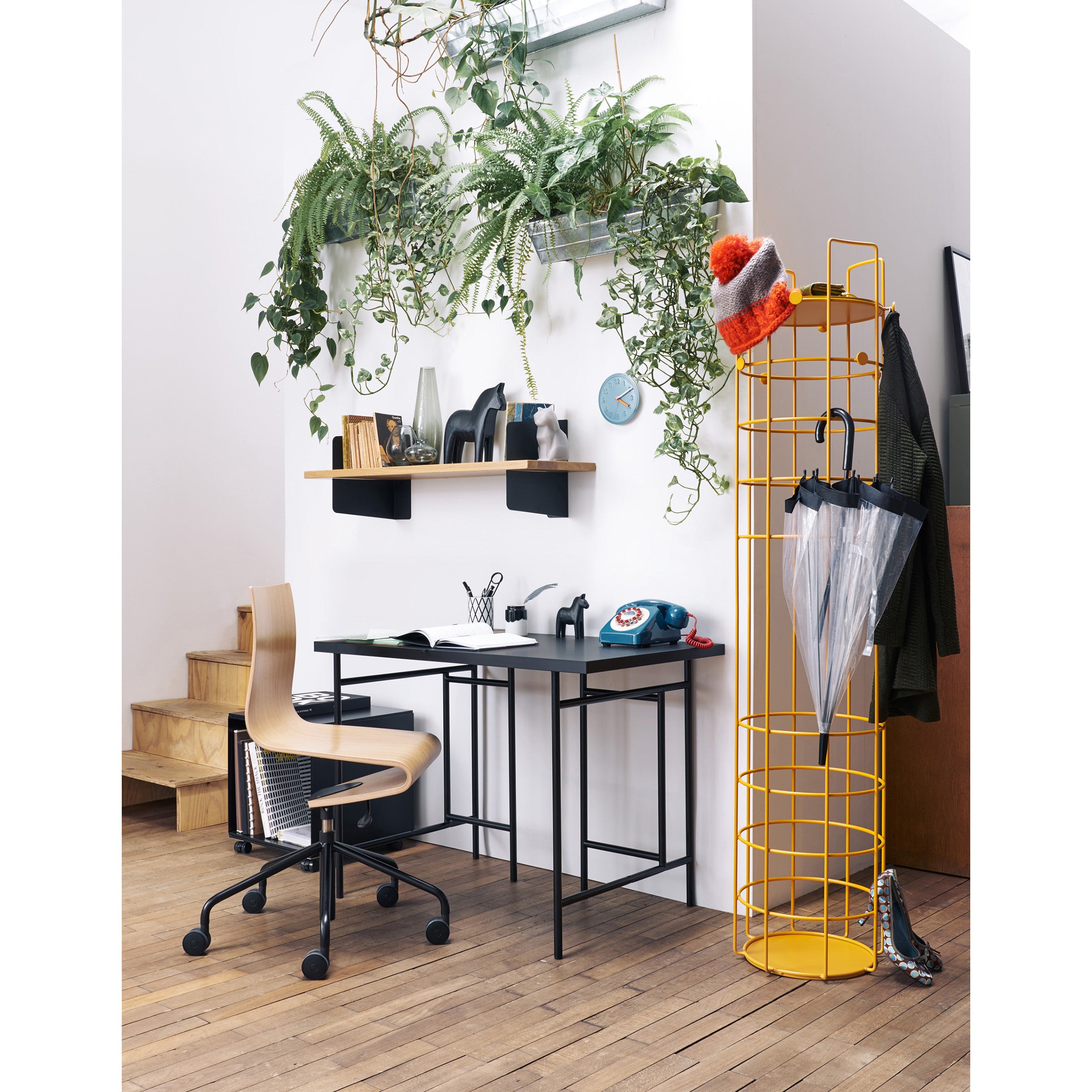 A modern workspace includes a light wood chair with wheels, a black desk, and the Richard Lampert Bazar Coat Stand for hats and umbrellas. A circular shelf with plants is mounted on a white wall under a skylight, while natural light brightens the wooden floor.
