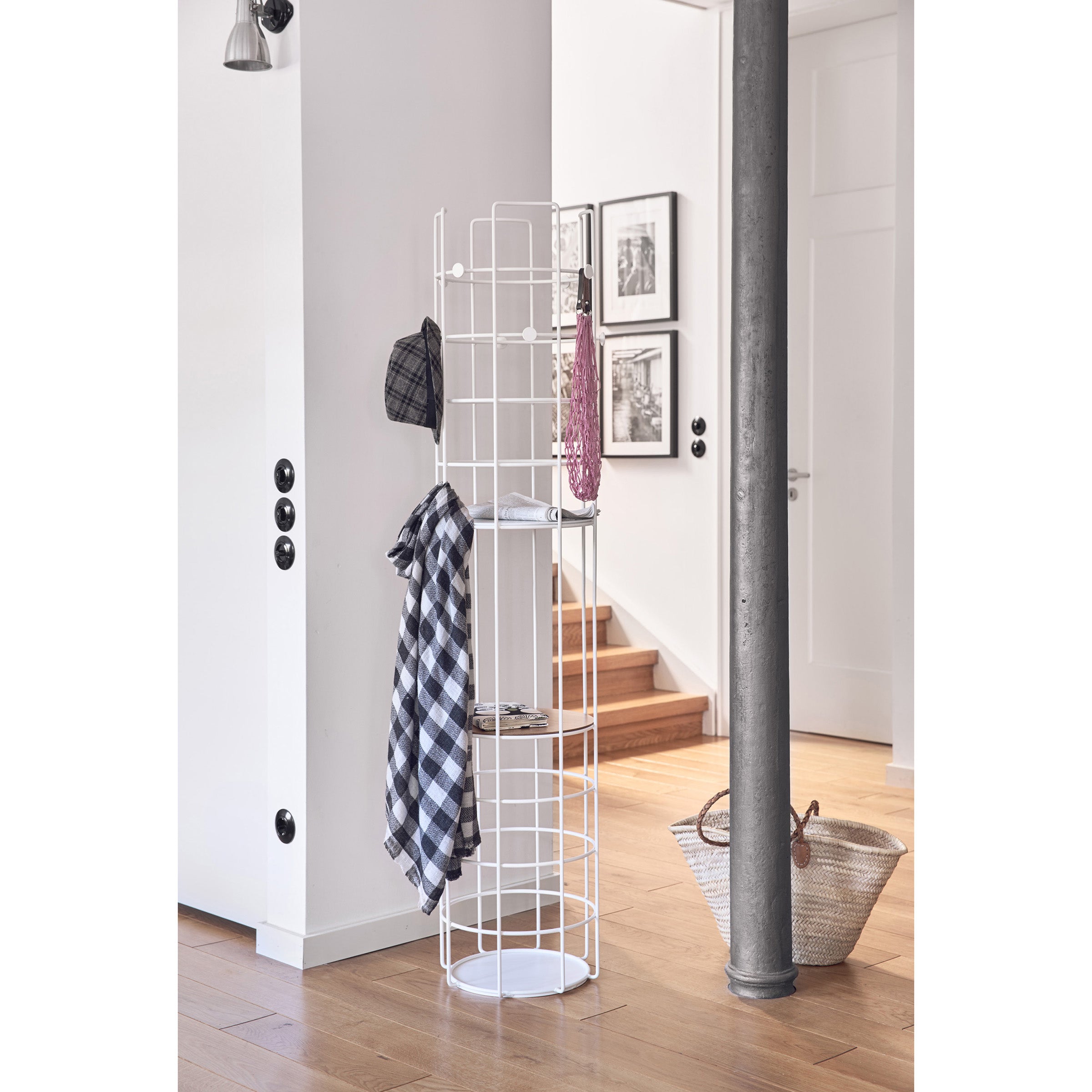 A modern interior highlights a Richard Lampert Bazar Coat Stand in white, holding a plaid shirt, hat, and bag. Nearby are framed pictures, stairs ascending, and a wicker basket on the wooden floor. A metal column and round light switches complete this practical setup.