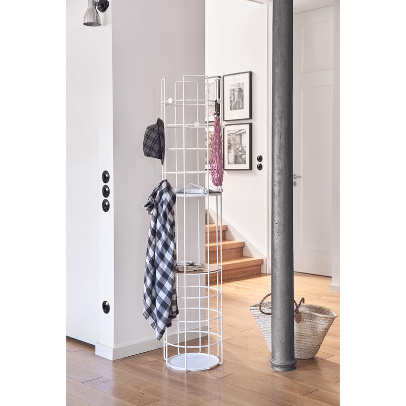 A modern interior highlights a Richard Lampert Bazar Coat Stand in white, holding a plaid shirt, hat, and bag. Nearby are framed pictures, stairs ascending, and a wicker basket on the wooden floor. A metal column and round light switches complete this practical setup.